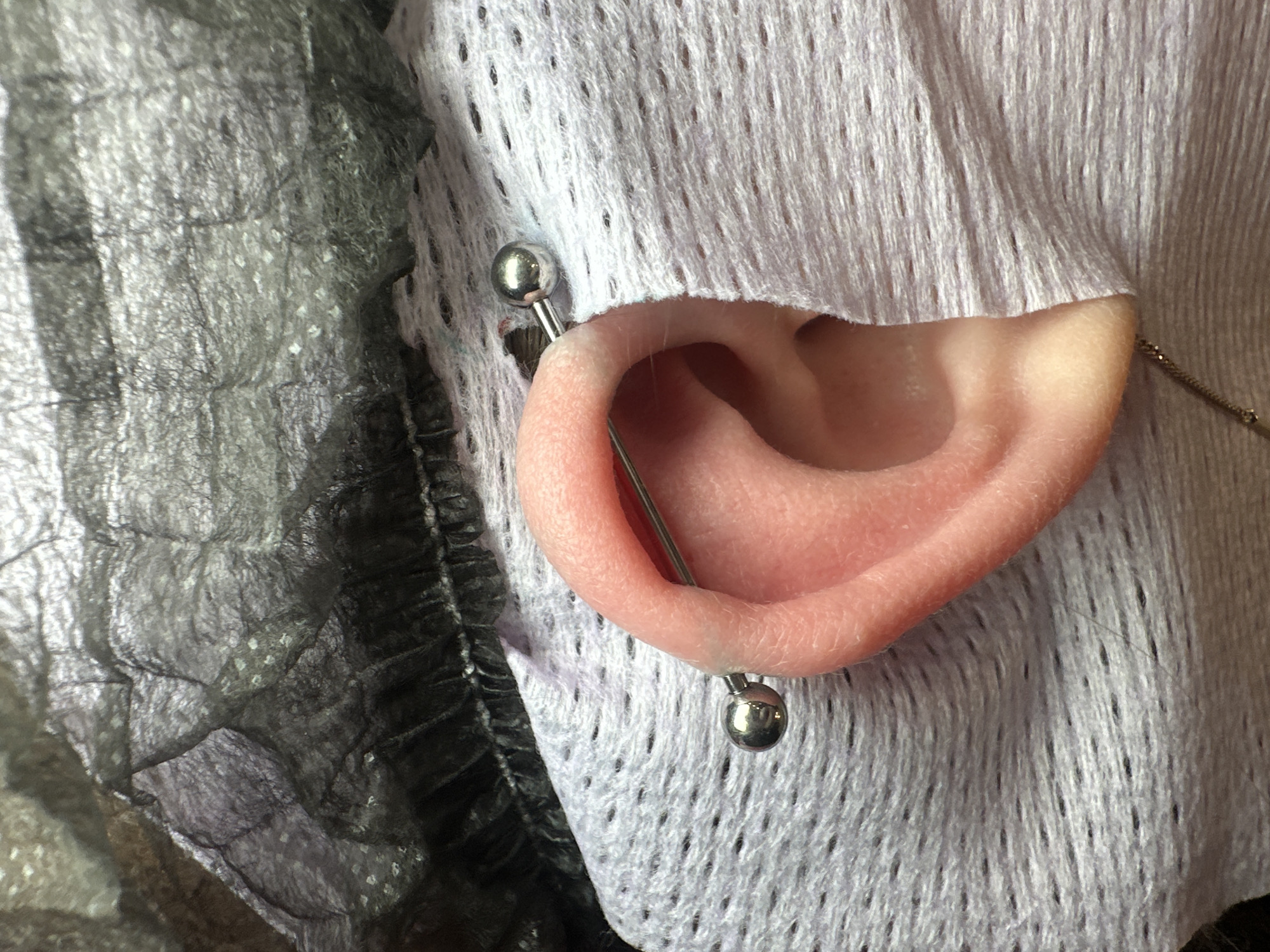 A close-up image shows a human ear with an industrial piercing. The piercing consists of a straight barbell passing through two separate piercings in the upper ear cartilage. The barbell has a small, silver ball on each end. The ear is pink and appears to be clean. The ear is positioned in front of two contrasting fabrics: a grey, crinkled fabric on the left and a white, perforated fabric on the right. The white fabric obscures part of the ear. A delicate chain is visible just behind the ear, near the back of the head.
The location is not provided by the image itself.