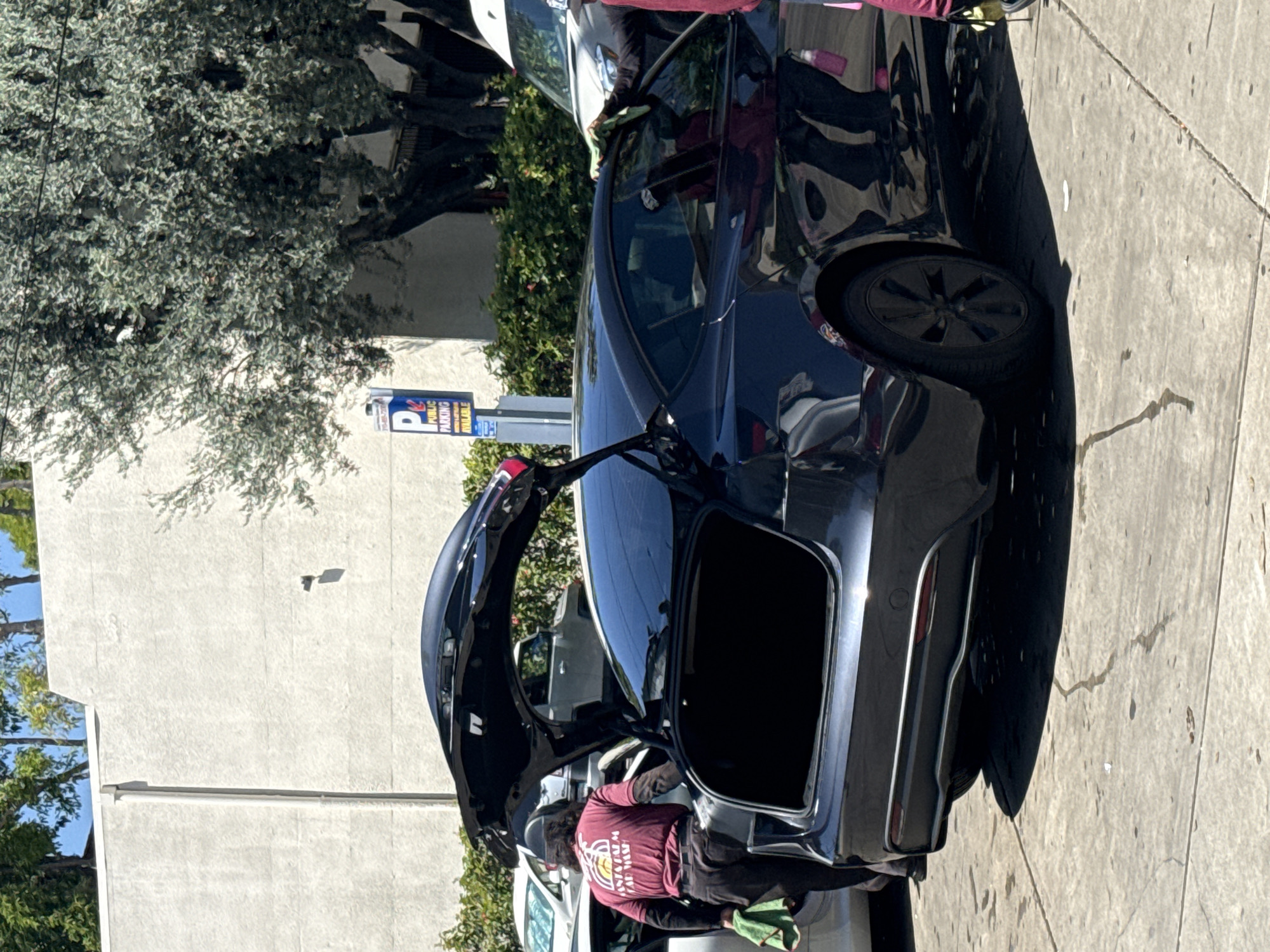The image shows a black Tesla Model X with its rear hatch open, parked on a sidewalk in West Hollywood, California. A person wearing a maroon shirt with a logo is leaning into the trunk, possibly cleaning or organizing items inside. The car is parked on the right side of the frame, adjacent to the sidewalk, with a building and trees visible in the background. A parking sign is also visible near the Tesla.