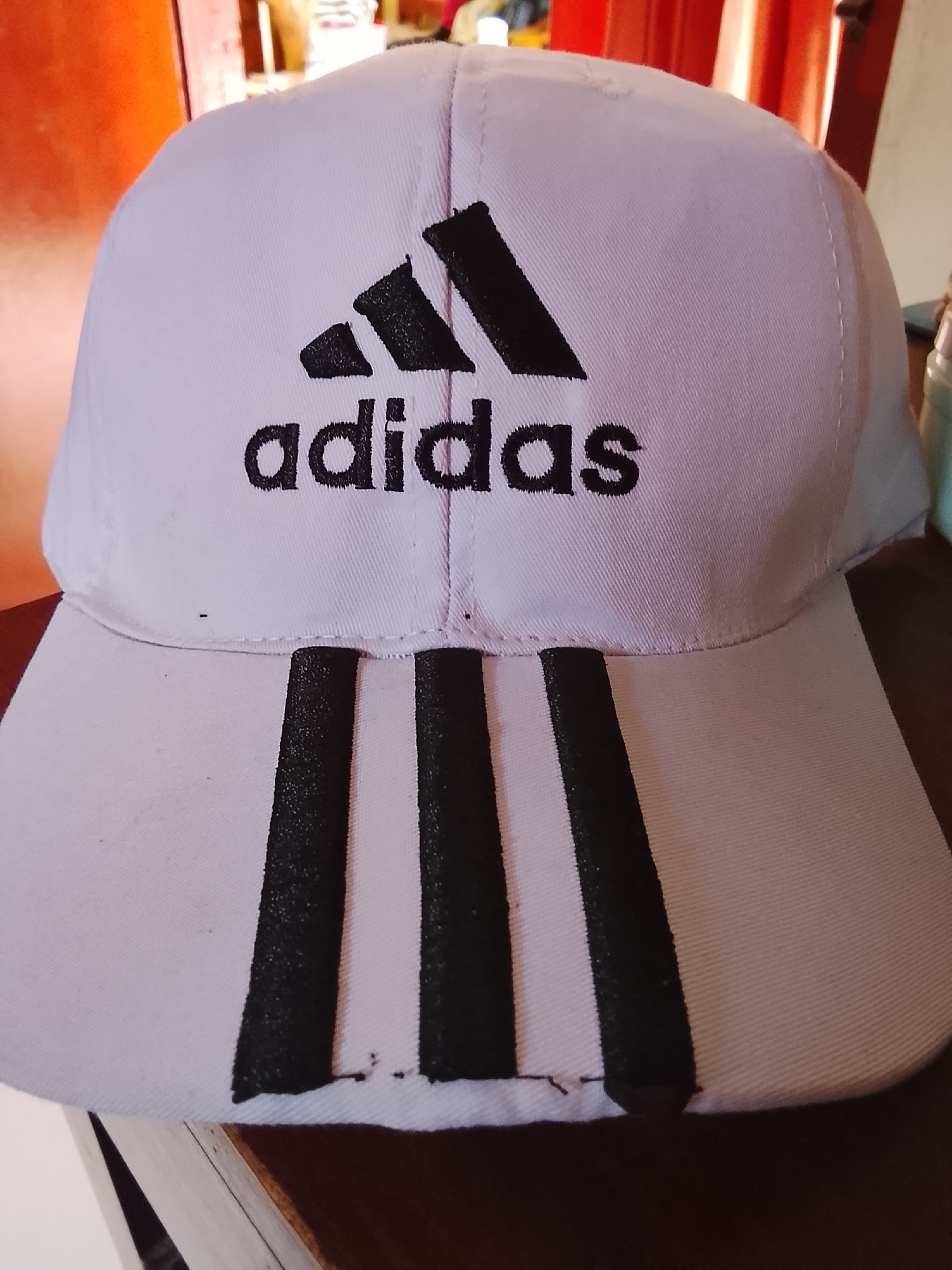 A close-up, eye-level, medium shot captures a white Adidas baseball cap with black lettering and three black stripes on the brim, displayed indoors on a dark wooden surface. The cap is positioned centrally, facing forward, with its brim slightly angled downwards. The background is a blur of orange and reddish hues, suggesting a wall or curtain. The lighting appears to be natural, coming from an unseen source. The image was taken in Karangklesem, Indonesia.
