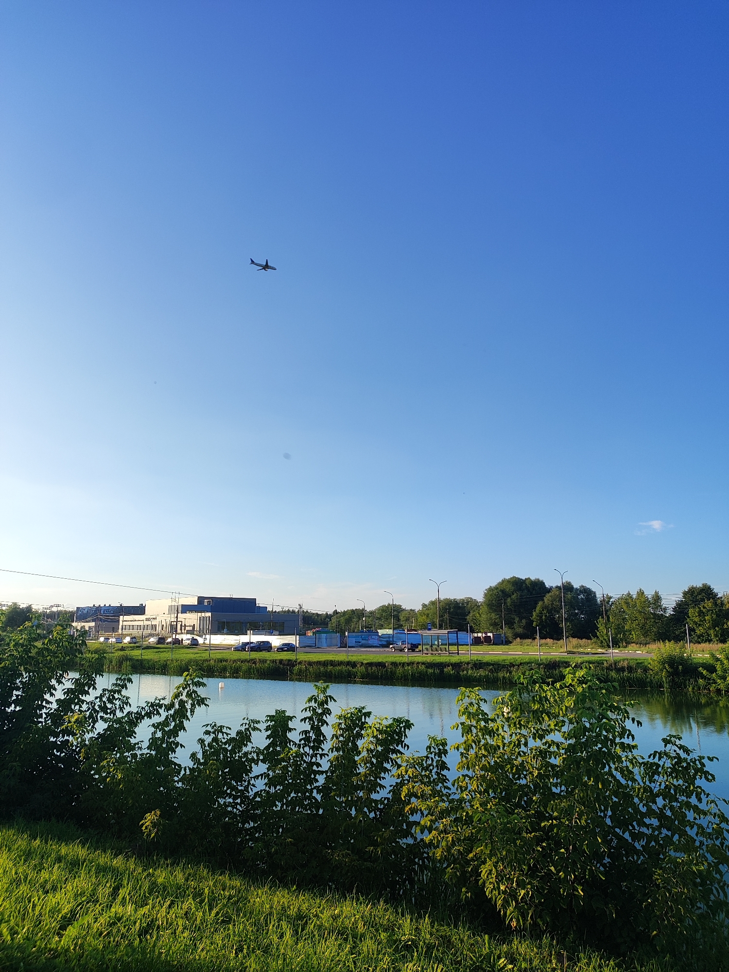 An aerial view of a bright blue sky over Bronnitsy, Russia, showcases a serene lake in the foreground, bordered by lush green grass and bushes. In the mid-ground, a modern industrial building with glass windows and a blue facade stands opposite a road lined with trees and streetlights. A small airplane can be seen flying high in the sky, adding a dynamic element to the otherwise tranquil landscape. The sun casts a warm glow on the scene, hinting at a pleasant summer day.