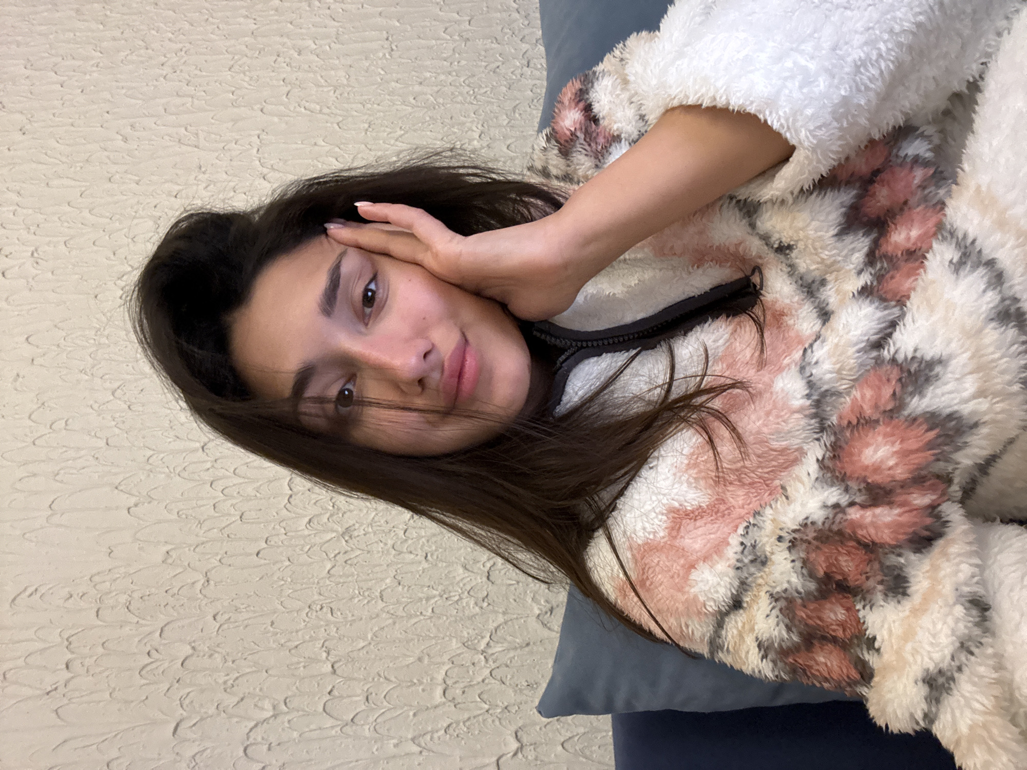 An individual is depicted indoors, seated on a dark blue sofa with a matching throw pillow to their left. The background consists of a light-colored, textured wall exhibiting vertical, undulating patterns. The subject is a female with long, dark brown hair, parted on the left side, with some strands partially obscuring her face. She has brown eyes and is looking directly at the viewer with a slight smile. Her left hand is positioned against the left side of her face, with fingers extended and resting near her temple and cheekbone. Her fingernails are light-colored. She is wearing a fuzzy, high-collared garment featuring an abstract pattern in white, light pink, and reddish-brown tones, with a visible black zipper partially open at the collar. The scene takes place in Sutomore, Montenegro.