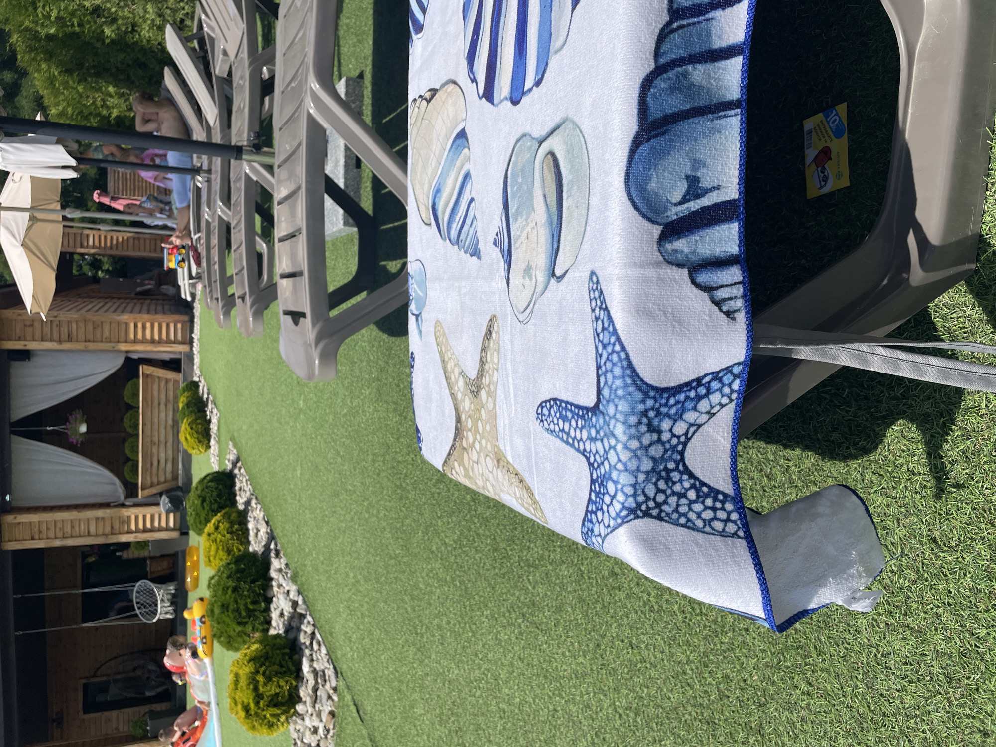 The image depicts a sunny outdoor scene at a recreational area, likely a resort or a private backyard, in Bohdanivka, Ukraine. In the foreground, a towel with a marine-themed print of seashells and starfish is draped over a lounge chair. The towel exhibits blue and white patterns. Behind the lounge chair, other identical chairs are arranged in rows, some occupied by individuals. To the left, a swimming pool is visible with children playing, some with inflatable toys. Further back, a wooden structure with a thatched roof and curtains suggests a cabana or gazebo. The ground is covered with artificial turf, interspersed with gravel and decorative bushes. The overall atmosphere is one of leisure and relaxation in a warm climate.