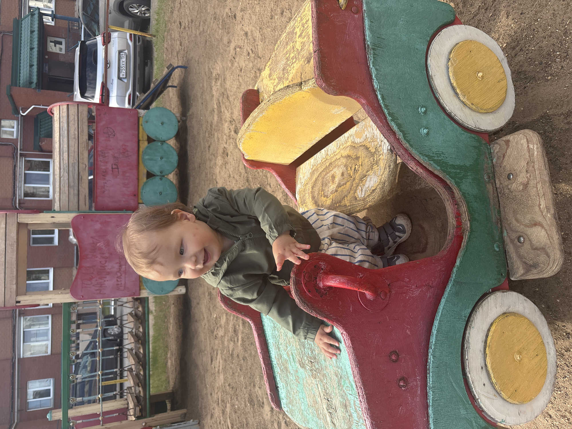 A cheerful young child is captured playing outdoors on a bright, sunny day in what appears to be a playground within a residential area. The child, with light hair and a happy, smiling expression, is positioned inside a colorful, weathered wooden play structure that resembles a car or part of a train. The structure is painted in vibrant, chipped red, green, and yellow, showing significant signs of wear from play. The child wears a green jacket and striped pants, with their small hands resting on the edges of the play structure.
The ground around the playground is sandy or dirt-covered. In the background, multi-story apartment buildings with numerous windows are visible, suggesting an urban or suburban setting. Several cars are parked near the buildings; the rear of a silver SUV shows a partially visible license plate reading "H755XX 152". To the left, another section of the playground, possibly a wooden train carriage, is painted red with three circular green elements, resembling wheels or traffic signals.
Faint, faded Cyrillic text, possibly graffiti, is visible on a red panel of the playground equipment behind the child's head. Some discernible words include "НАИВНЫЕ" (NAIVNYE) and "МЕГА" (MEGA). The overall scene conveys a sense of innocent play and joy in a typical outdoor community setting in Chaikovsky, Russia.