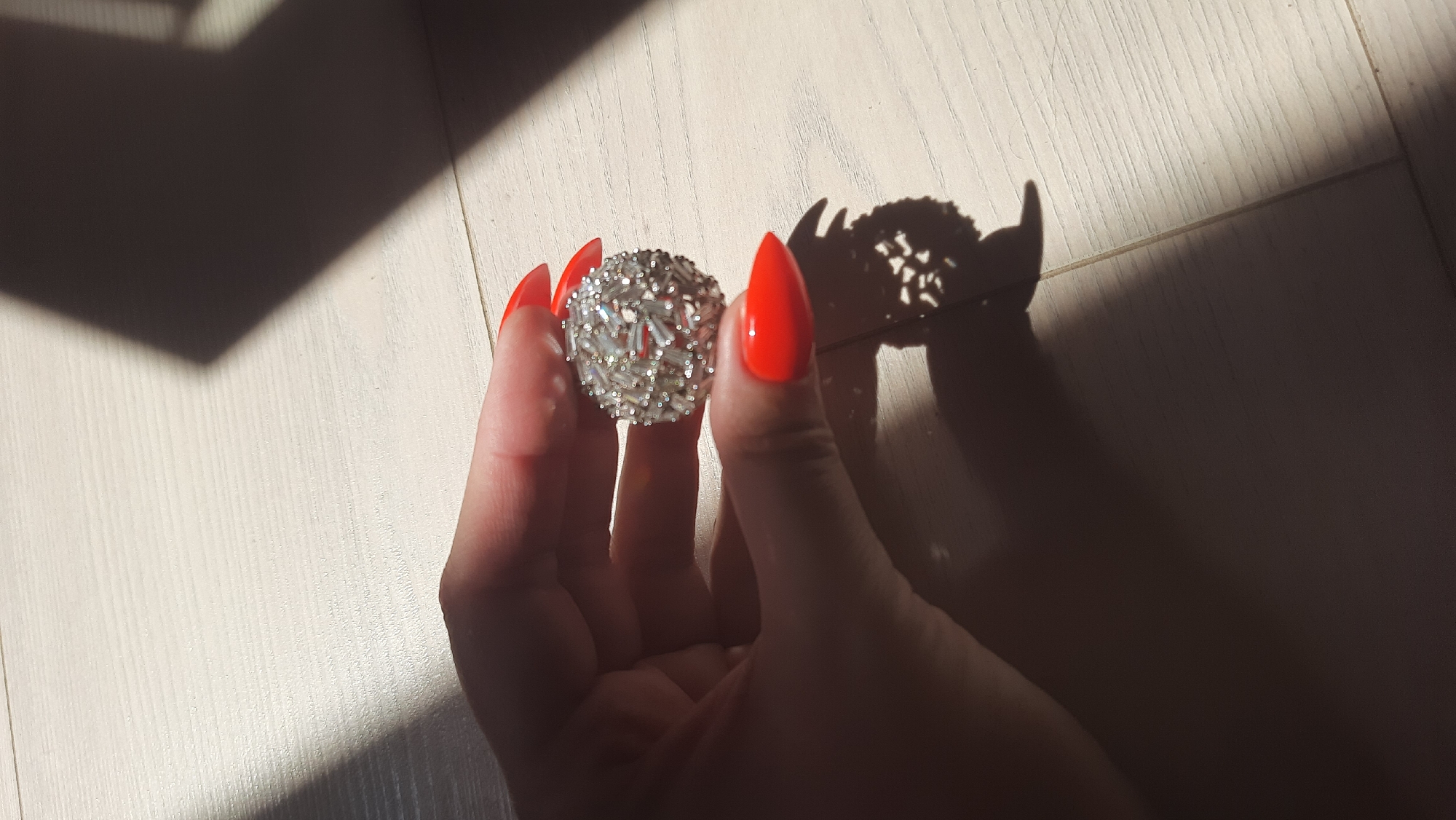 A person's hand with bright orange, stiletto-shaped fingernails holds a round, sparkling ring composed of numerous small, baguette-cut stones. The ring is being held in front of a light-colored wooden surface, likely a floor, which is illuminated by diagonal shadows cast by sunlight. One shadow is large and abstract, falling across the background, while a smaller, more intricate shadow of the ring itself is visible on the surface to the right. The image is a close-up, focused on the hand and the ring. There is no information about the location in Desnogorsk, Russia.