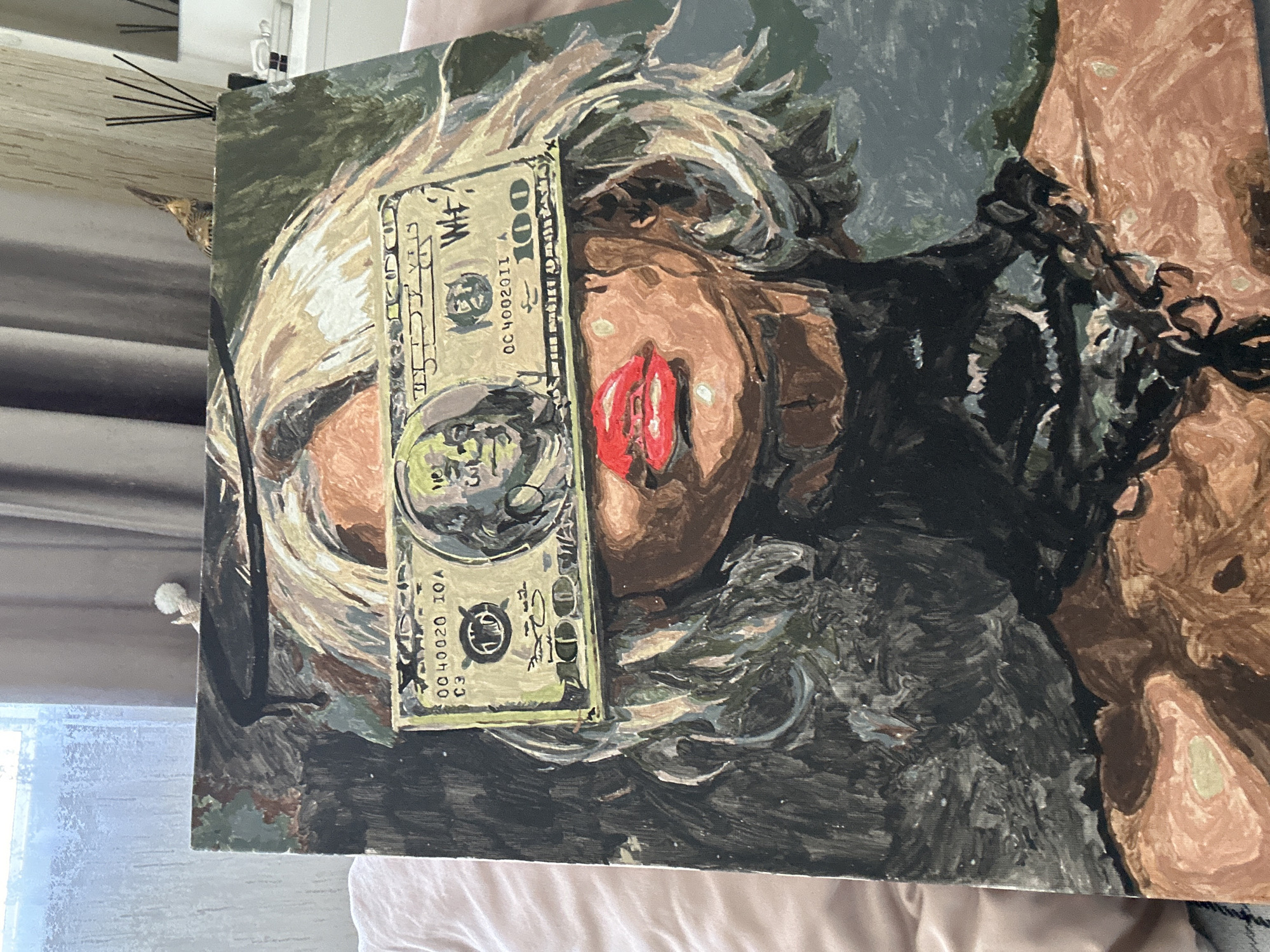 The image shows a painting of a woman. Her face is partially obscured by a $100 bill. The woman has light blonde hair, red lips, and brown skin. She appears to be wearing a dark outfit, with a chain around her shoulder. The background is a mix of greens and grays. The painting rests on a light pink surface. There is no evidence to indicate the location as Высокий, Russia.