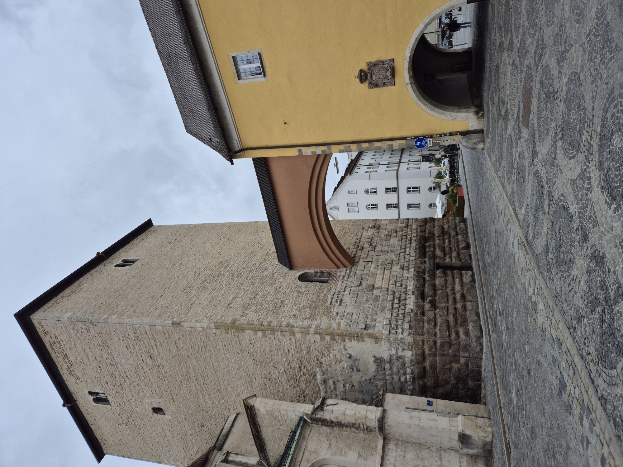 The image captures a view of the historic city of Regensburg, Germany, specifically focusing on a section of the old town. In the foreground, a cobblestone street winds its way between two significant architectural structures. To the left, a tall, imposing stone tower with small, dark windows rises against a cloudy sky. This appears to be part of a larger stone building, possibly a church or a fortification, with lighter-colored stone sections and a rougher, darker base.
Connecting the tall stone structure to a building on the right is a wide, arched walkway or bridge with a terracotta-colored roof and an orange-brown supporting arch.
To the right, a vibrant yellow building stands prominently. It features a single white-framed window near the top and a decorative crest with a crown above an arched entryway. This archway serves as a passage into the building or beyond. Inside the archway, the darkness suggests an interior space or further passage.
In the mid-ground, a white building with multiple windows is visible through the archway of the yellow structure. The street level in front of this white building shows tables and umbrellas, suggesting a possible outdoor seating area for a cafe or restaurant. People can be seen in the distance, walking and interacting in this area.
A blue traffic sign with a bicycle symbol and the word "Ende" (meaning "End" in German) is mounted on a pole near the yellow building, indicating a change in traffic rules or the end of a designated zone.
The overall atmosphere is overcast and muted, with a sense of historical depth conveyed by the ancient stonework and architecture. The scene depicts a typical European city street with a blend of historical landmarks and contemporary signs of life.
