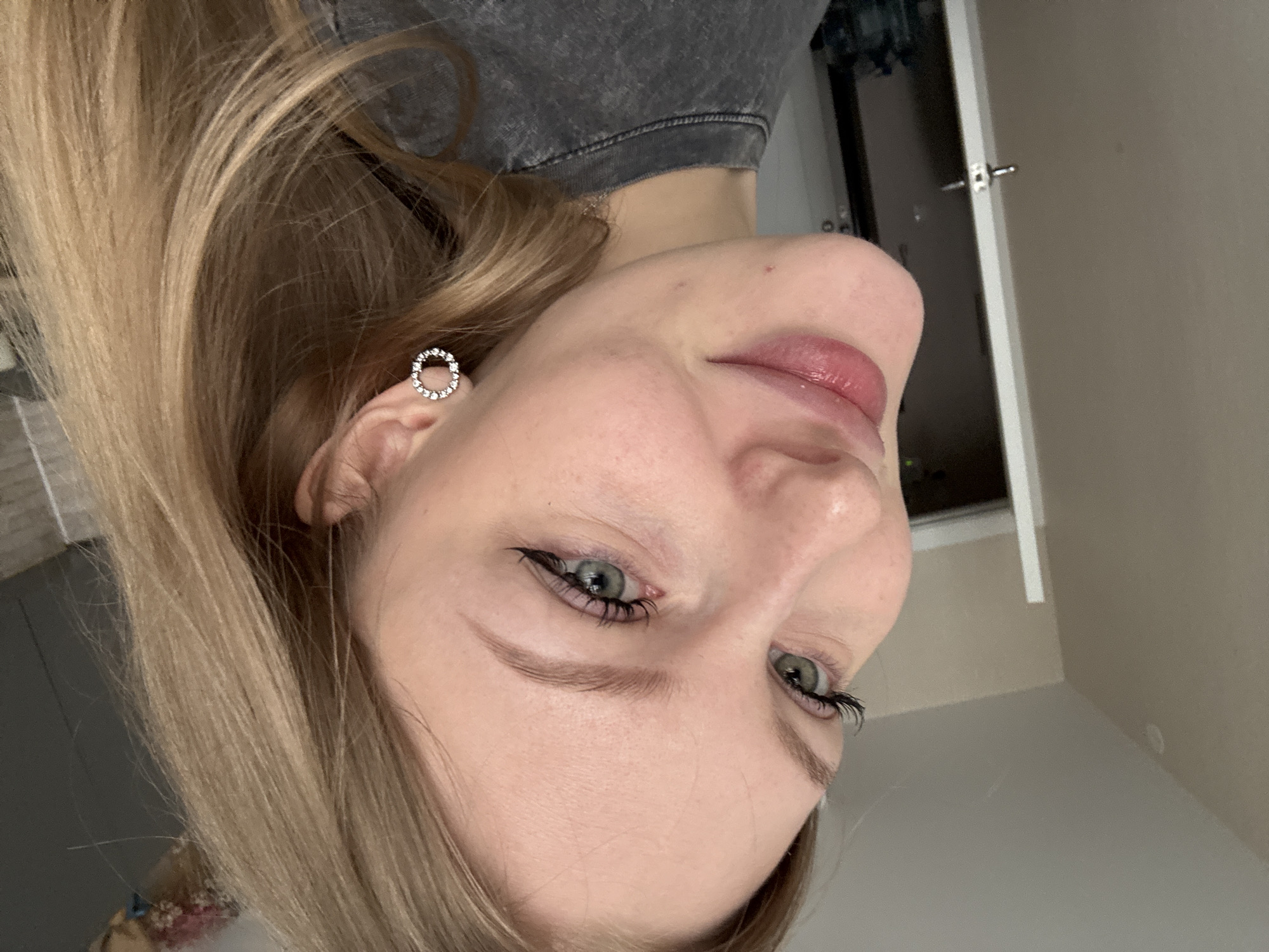A close-up, rotated portrait captures a fair-skinned person with light brown hair, wearing a gray top and small, circular earrings. The person has light eyes enhanced by black eyeliner and pinkish lips. The background shows a glimpse of a dark-colored object, a door, and a light-colored wall. No specific details indicate the city of Perm, Russia.