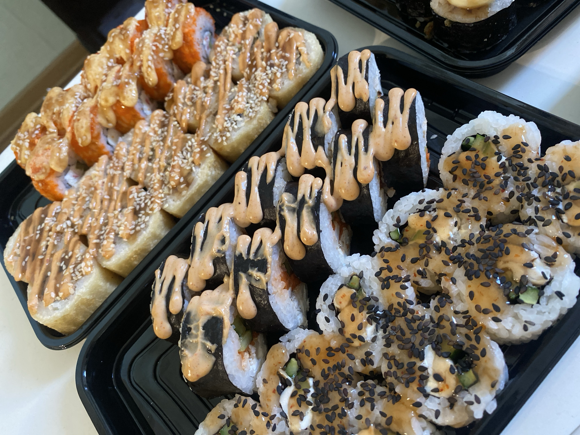 The image displays two black plastic take-out containers filled with various types of sushi rolls, positioned on a light-colored surface.
In the container on the left, several rows of sushi rolls are visible, featuring an outer layer with an orange hue. These rolls are generously drizzled with a light brown or tan creamy sauce and topped with white sesame seeds. Some of these rolls exhibit a slightly crispy texture, suggesting a potential tempura-style preparation.
The container on the right presents two distinct sushi styles. The upper portion contains rolls with dark green or black seaweed (nori) on the outside, also covered with the same light brown/tan creamy sauce. The lower section of this container holds uramaki-style rolls, characterized by white rice on the exterior. These rolls reveal fillings such as green cucumber and a white, creamy substance, and are heavily garnished with a glossy sauce and numerous black sesame seeds.
The sushi rolls are neatly arranged in rows within their containers, indicative of prepared food from a restaurant or a take-out order. The scene is brightly and evenly lit, suggesting an indoor setting during the day. No text is visible in the media file. The location context for this image is Sochi, Russia.