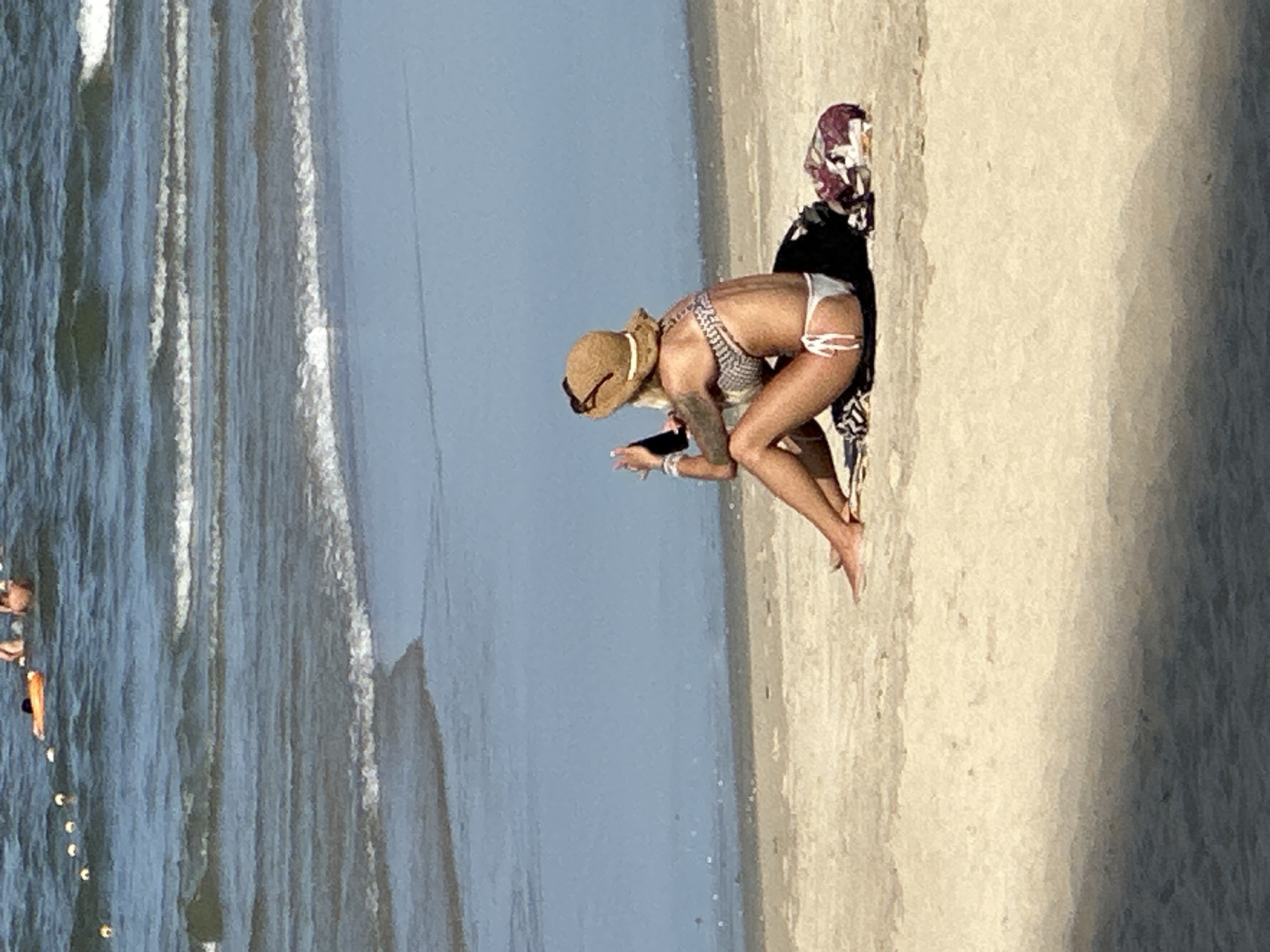 A woman in a straw hat and a bikini sits on a sandy beach in Da Nang, Vietnam, holding a smartphone. She is facing away from the camera, with her legs crossed. A patterned towel or blanket is behind her. The ocean is visible in the background, with gentle waves breaking on the shore. In the distance, several people are in the water, some with an orange float. The overall scene depicts a relaxed day at the beach.