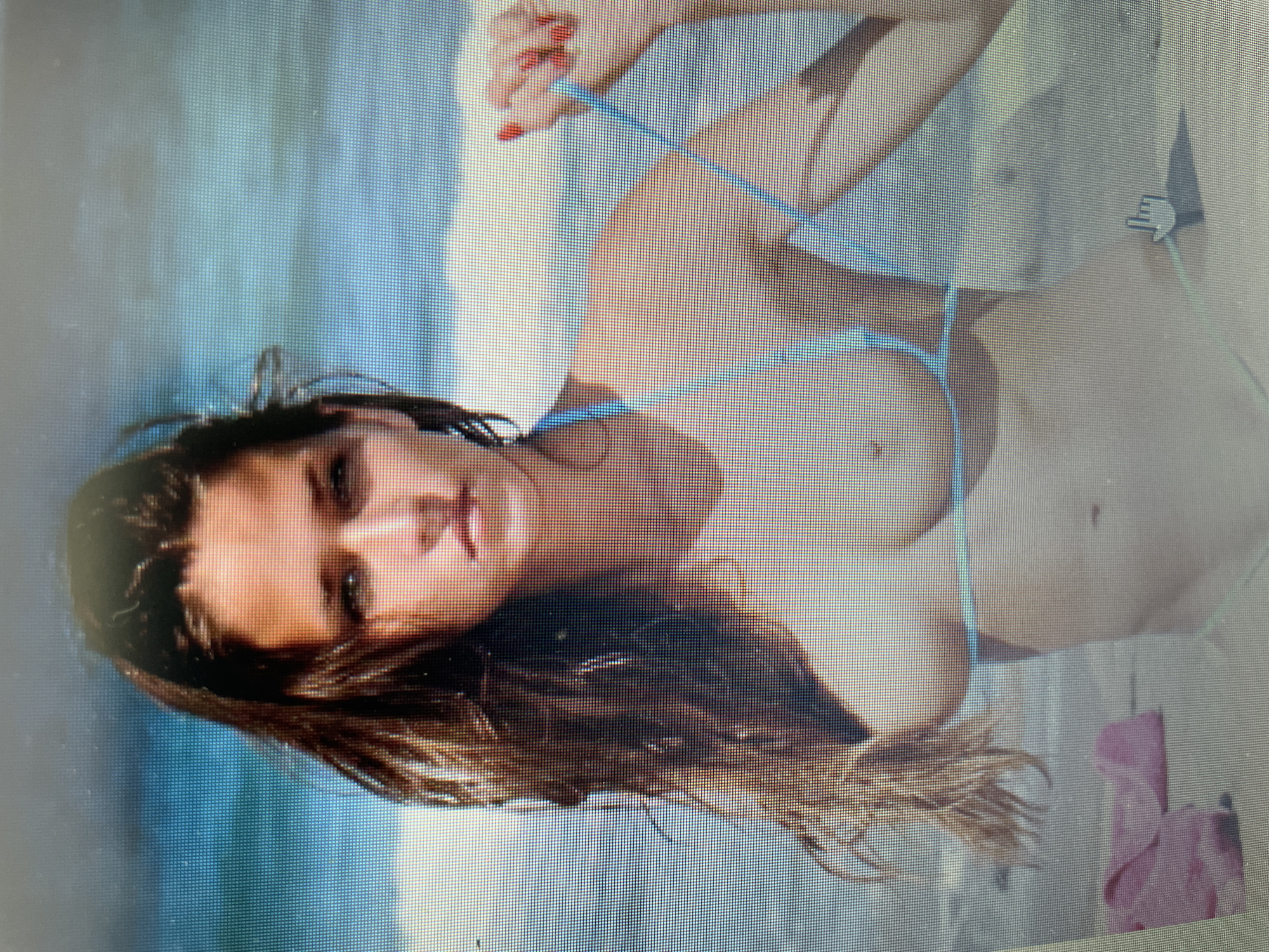 This media file depicts a portrait of a woman lying down on a beach. She has long, dark hair and is wearing a blue bikini. Her eyes are open and she is looking towards the camera. The background appears to be the ocean with some waves. The lighting suggests it is daytime, and the setting is a beach. There are no visible text elements in the image. The overall mood is relaxed and serene. The woman's pose is casual, and her expression is neutral. The image has a somewhat grainy quality, possibly due to the resolution or display it is being viewed on.