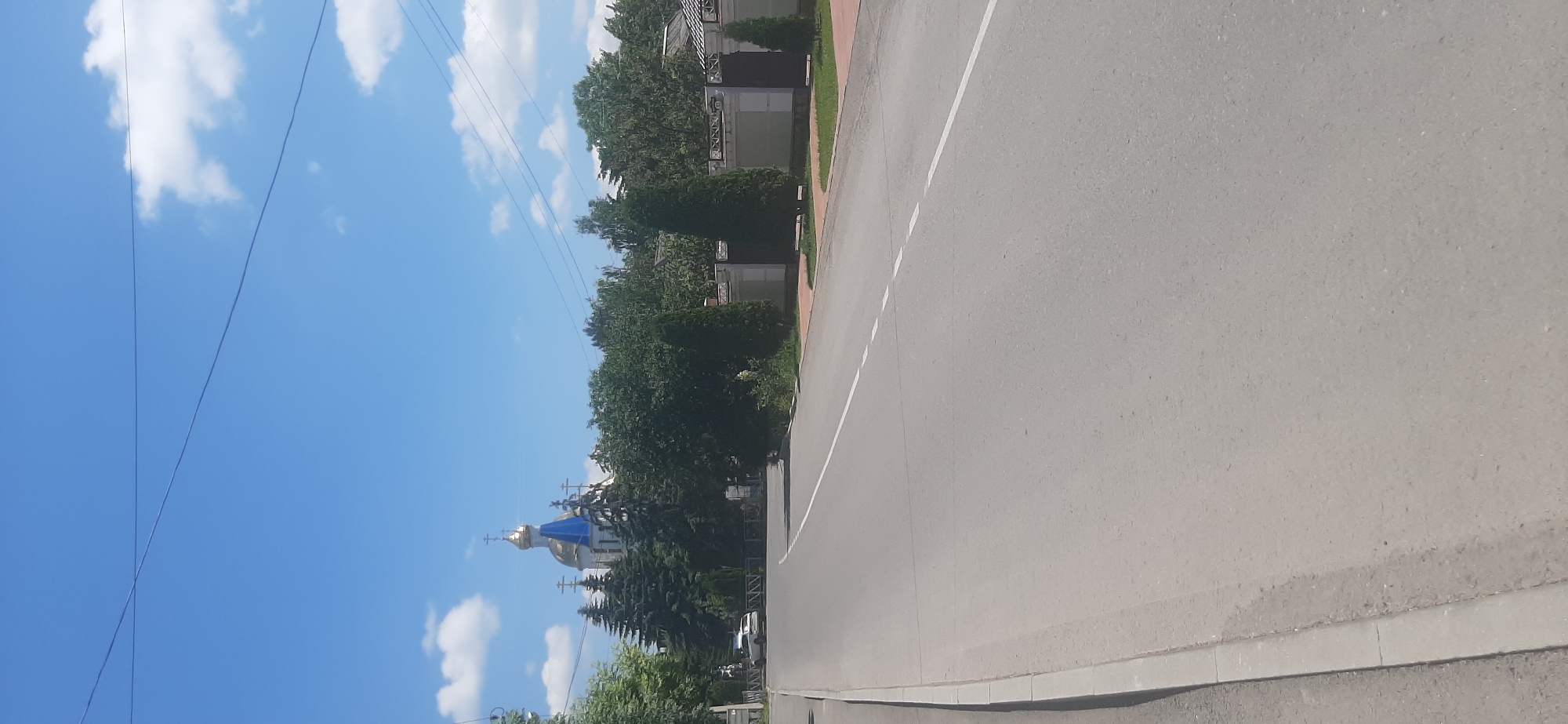 The image shows a street scene in Yessentukskaya, Russia. A paved road with white lane markings runs through the center of the frame, with a sidewalk on the left. On the right side of the road, there is a white fence with dark gates, behind which are trees and the roof of a building. In the background, a church with golden domes and a blue spire is visible, surrounded by evergreen trees. A white car is parked on the side of the road to the left of the church. The sky is blue with scattered white clouds. The overall impression is of a sunny day in a town or village.