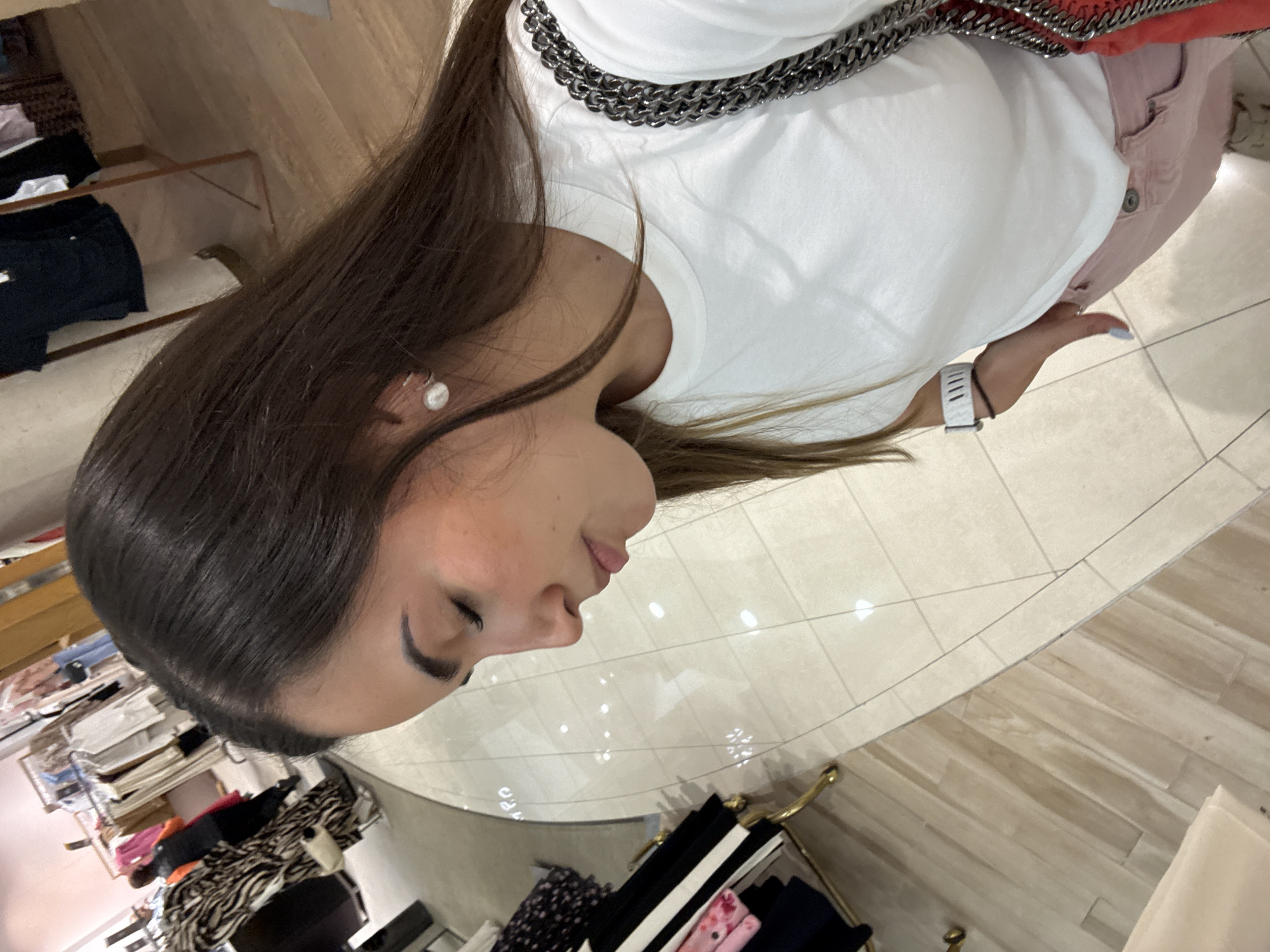A woman is photographed from above inside what appears to be a retail establishment in Puebla, Mexico. She wears a white t-shirt with a chain detail around the neck, pink pants, and a white wristwatch. Racks of clothing are visible in the background, indicating a store environment. The flooring is tiled, further suggesting a retail space.