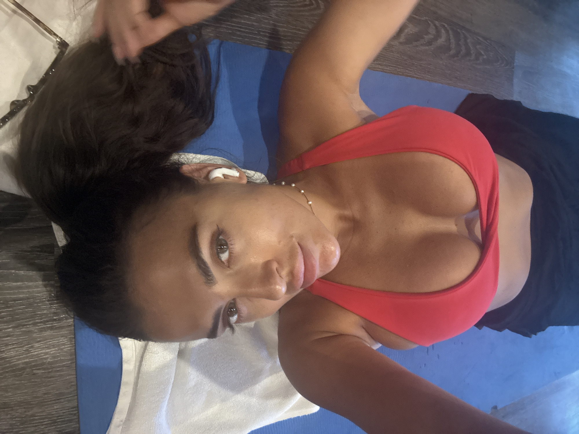A person with dark hair is lying on a blue yoga mat. She is wearing a red sports bra and black shorts. A white towel is next to her on the mat. The person is looking at the camera. There's an object near her hair that is potentially a hairbrush. There is no evidence that this image was taken in North Kuta, Indonesia.