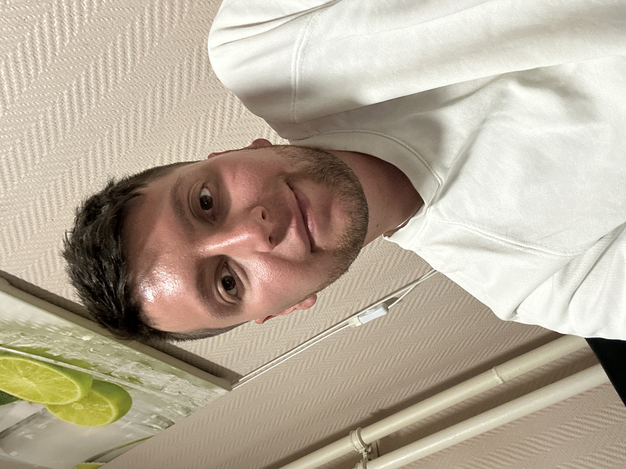 A man is looking down at the camera. He is wearing a white sweatshirt. Part of a lime-themed picture is visible in the bottom left corner. The location is a room with beige wallpaper and white pipes. There is no evidence to suggest this image was taken in Vorotynsk, Russia.