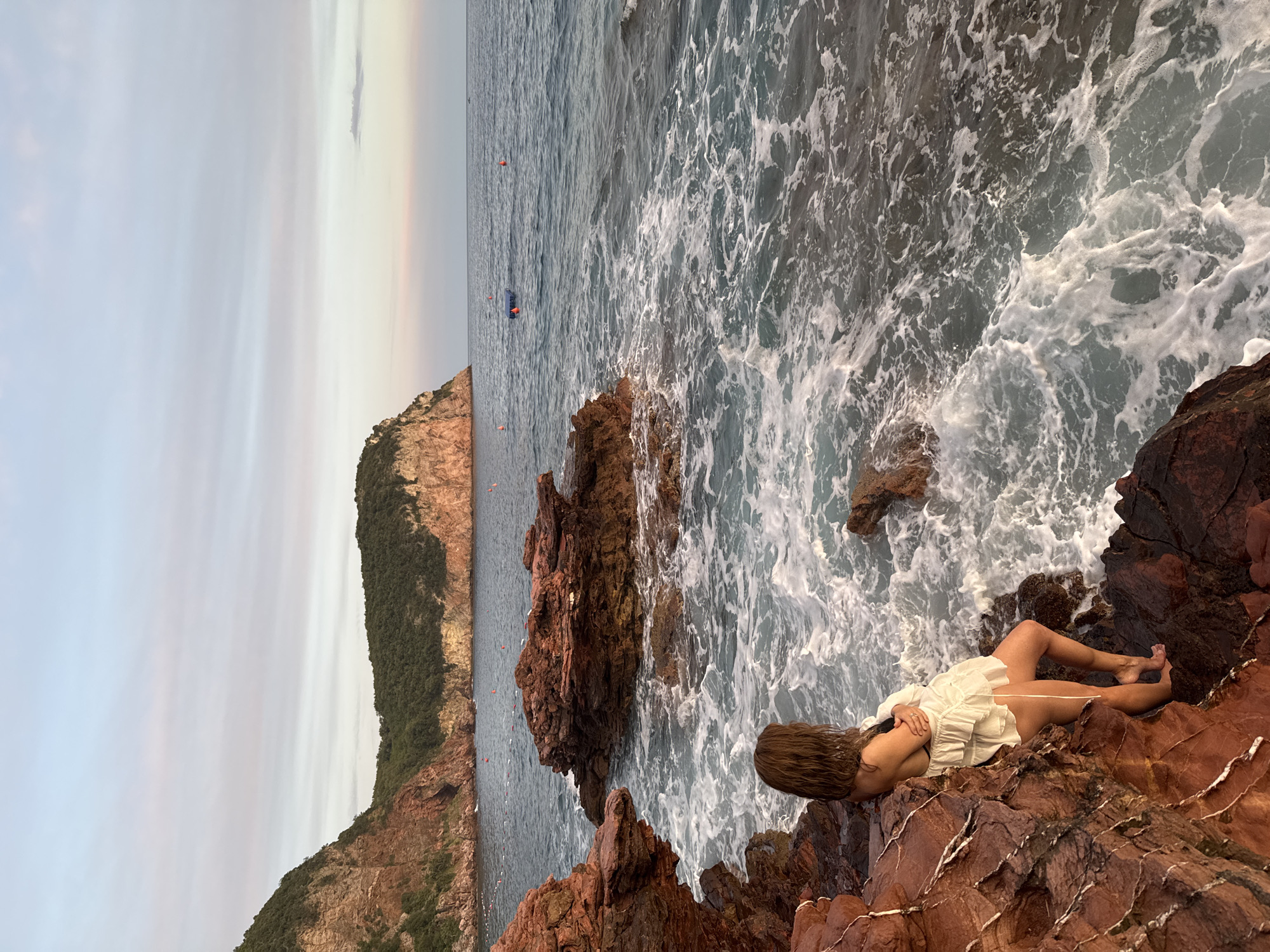 A woman with long brown hair, wearing a white ruffled dress and a black bikini top, stands on reddish-brown rocks near the sea in Sveti Stefan, Montenegro. She is looking out at the water, with her back to the viewer. The sea is choppy with white foam crashing against the rocks. In the background, a rugged coastline with green vegetation and cliffs is visible. The sky is a soft gradient of pale blue and pink, indicating either sunrise or sunset. Several orange buoys and a blue floating platform are scattered in the water.