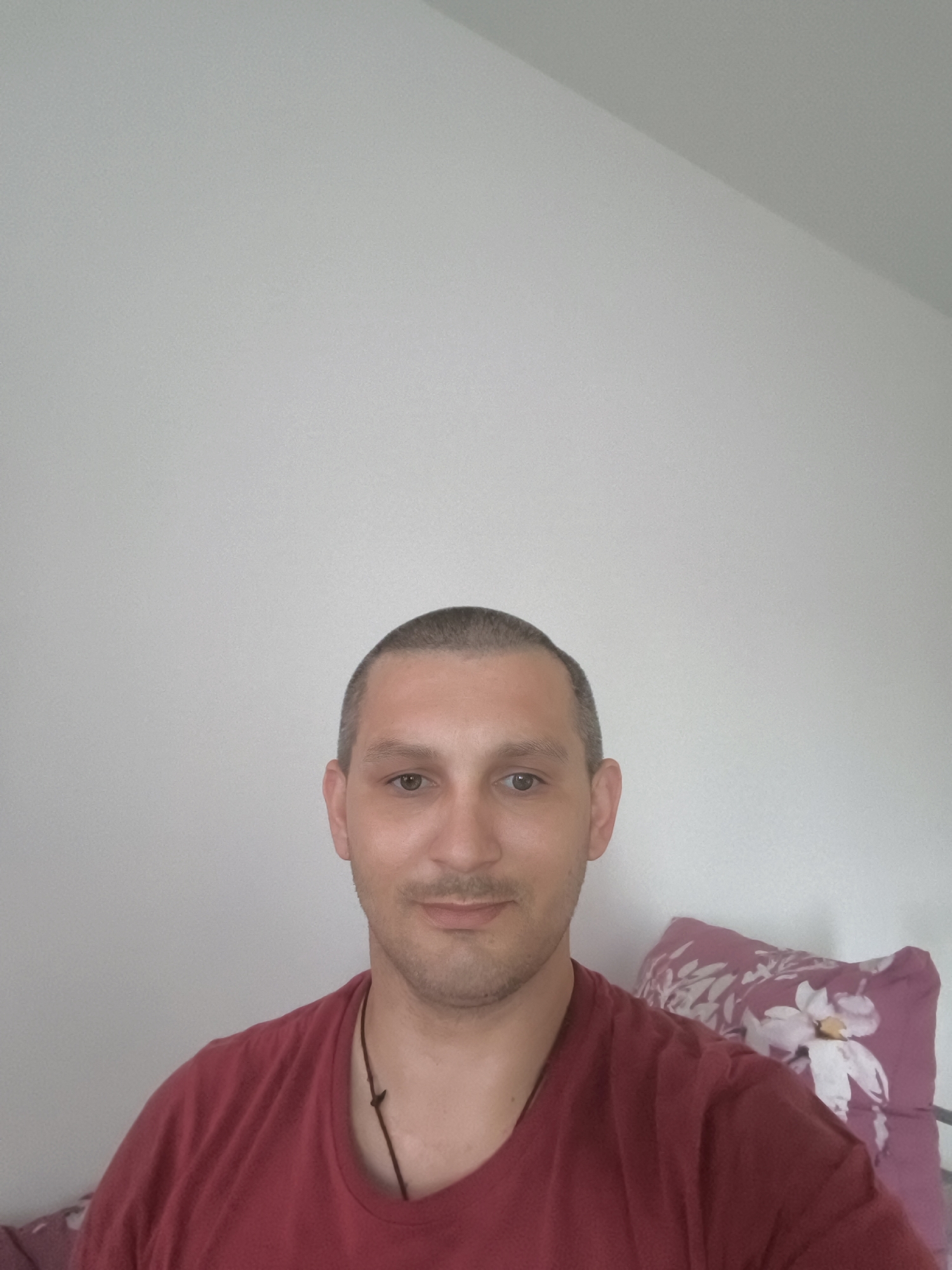 A man with a shaved head and a light mustache is looking directly at the camera. He is wearing a dark red t-shirt with a thin cord necklace. Behind him, a pillow with a floral pattern is visible. The background is a plain, light-colored wall. There is no information in the image to suggest a location in the city of Wolgast, Germany.