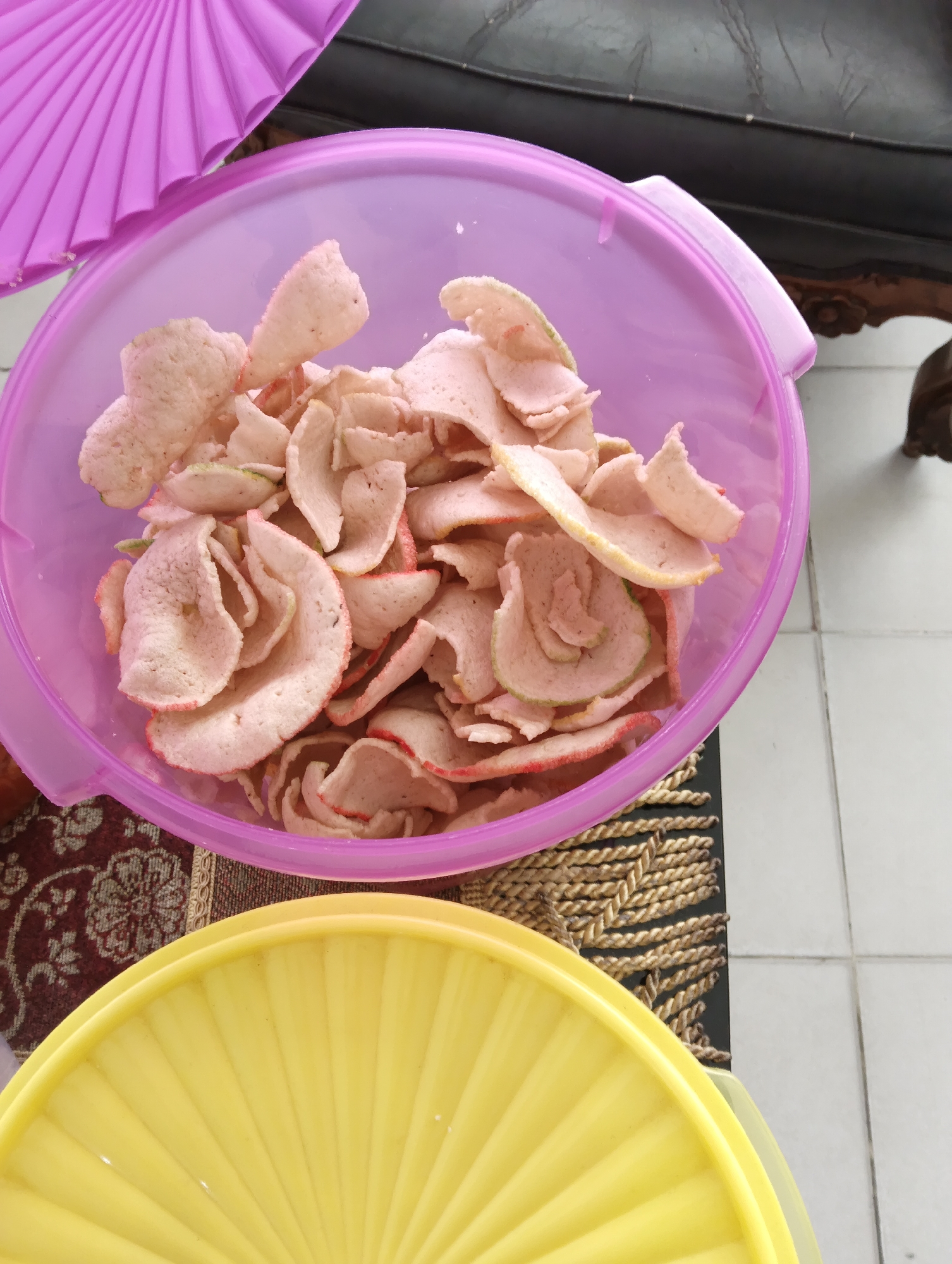 This is a close-up, eye-level, indoor shot of a pink plastic bowl filled with pale pink crackers with red edges. The bowl is on a dark table with a patterned brown placemat. To the left of the bowl, a portion of a purple fan-shaped object is visible. In the foreground, a bright yellow plastic lid with a ribbed pattern is partially visible. The background shows a dark leather surface, possibly a sofa, and part of a tiled floor. The scene appears to be in a residential setting in Pekanbaru, Indonesia. There are no people visible.