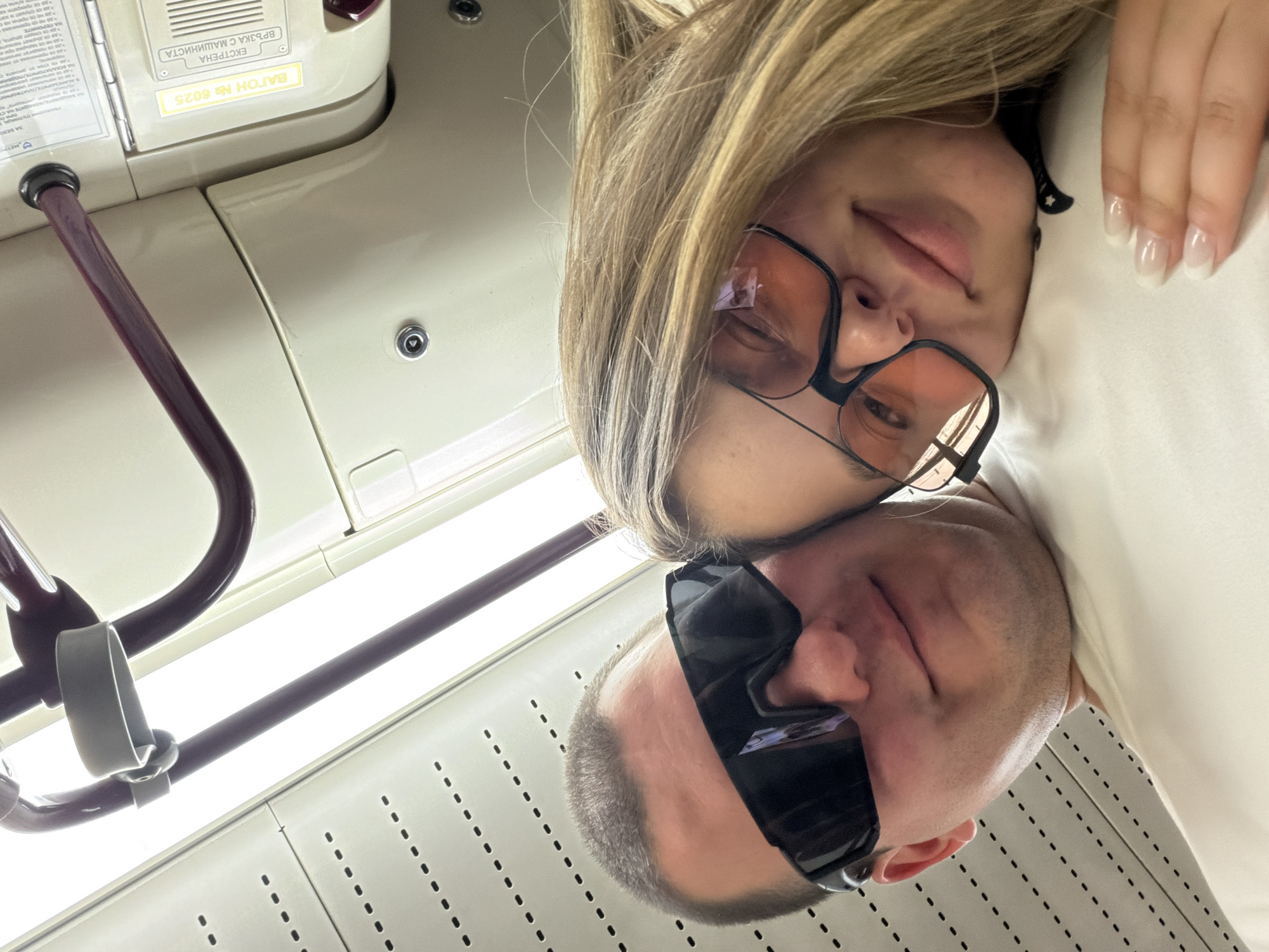 In a selfie taken from a low angle inside a public transport vehicle, likely a subway car in Sofia, Bulgaria, a couple is captured in a close embrace. The ceiling of the vehicle, with its textured panels and dotted pattern, is visible in the upper portion of the frame. The man, positioned behind and slightly above the woman, wears large, dark sunglasses and rests his head against hers. His facial features are partially obscured by the angle and his sunglasses.
The woman, in the foreground, is looking towards the camera with a slight smile and one eye closed in a wink. She also wears distinctive oversized glasses with light orange tinted lenses. Her blonde hair frames her face, and her left hand, adorned with long, pale fingernails, rests on the man's shoulder or chest. The interior of the subway car is visible in the background, with cream-colored panels and burgundy handrails. A yellow sign with Cyrillic text, "ВАГОН № 6025" (CAR No. 6025), and "ВРЪЗКА С МАШИНИСТА" (CONNECTION WITH THE DRIVER), is affixed to a panel, indicating the specific location within the train. The overall lighting suggests an indoor environment, possibly during daytime, with some glare from overhead lights. The scene conveys a sense of intimacy and casual travel.