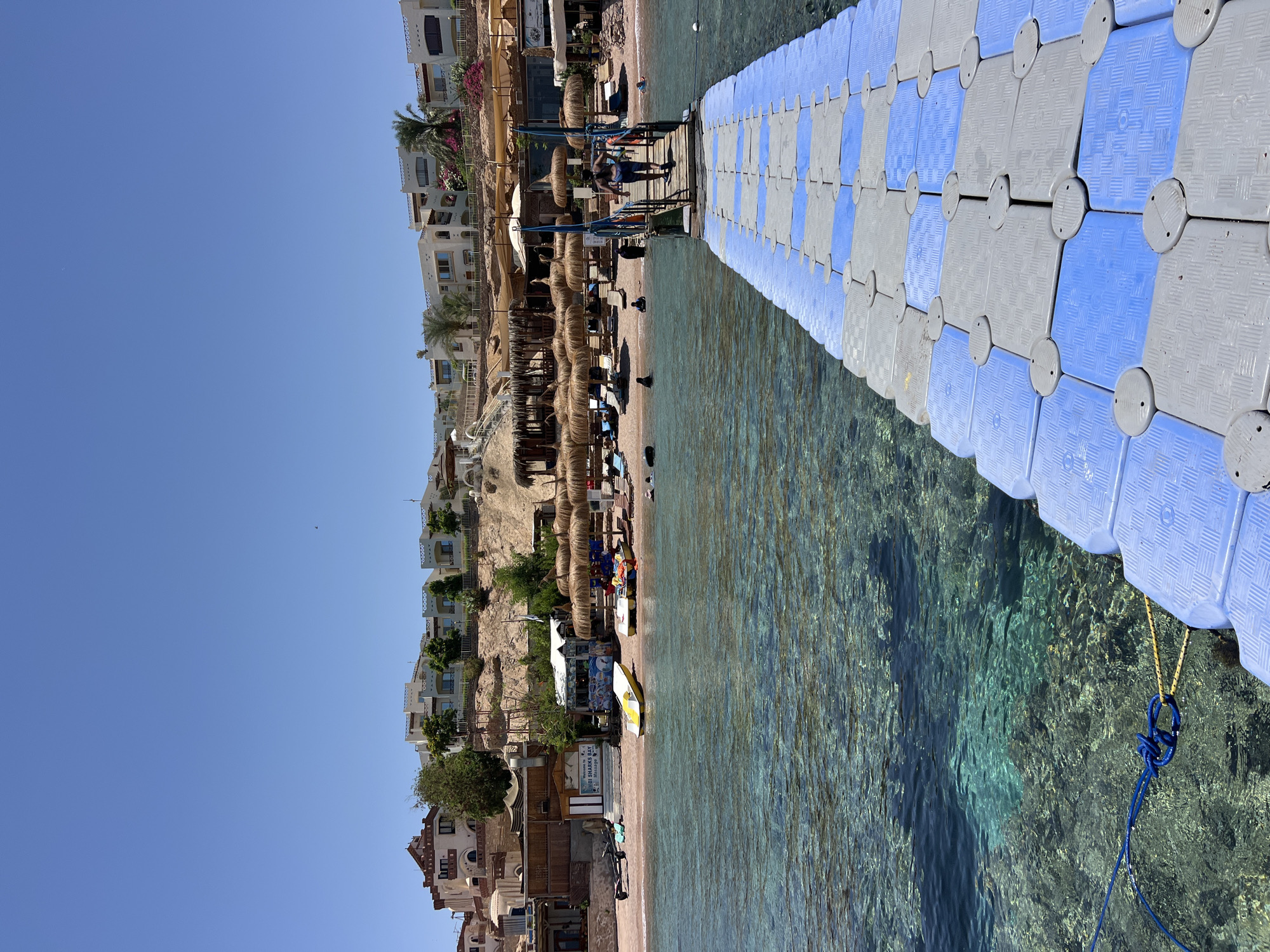 A vertical, eye-level shot shows a beach resort area in Sharm El Sheikh, Egypt. The scene is bathed in bright daylight, suggesting a sunny day with clear blue skies.
On the left, a hillside is dotted with white, multi-story buildings, likely hotels or apartments, with balconies and windows visible. The buildings are nestled into the rocky terrain, with some greenery and palm trees interspersed. Below the buildings, a beach stretches out, lined with numerous thatched-roof sun umbrellas and lounge chairs. People are visible on the beach, some relaxing under the umbrellas, others walking or wading in the shallow water.
To the right, a floating pontoon walkway extends into the clear, turquoise water. The pontoon is made of interconnected modular plastic blocks, alternating in blue and grey. The water is exceptionally clear, allowing the rocky seabed to be seen beneath the surface. The water appears calm, with gentle ripples on its surface.
A blue rope is tied to the pontoon and trails into the water. In the foreground, the pontoon takes up a significant portion of the frame. The beach area appears to be a vibrant hub of activity, with beach umbrellas, a small stand or kiosk, and beachgoers. The overall mood is relaxed and inviting, typical of a beach vacation destination.
Visible text includes "Welcome to NUBI SHARKS BAY Massage".