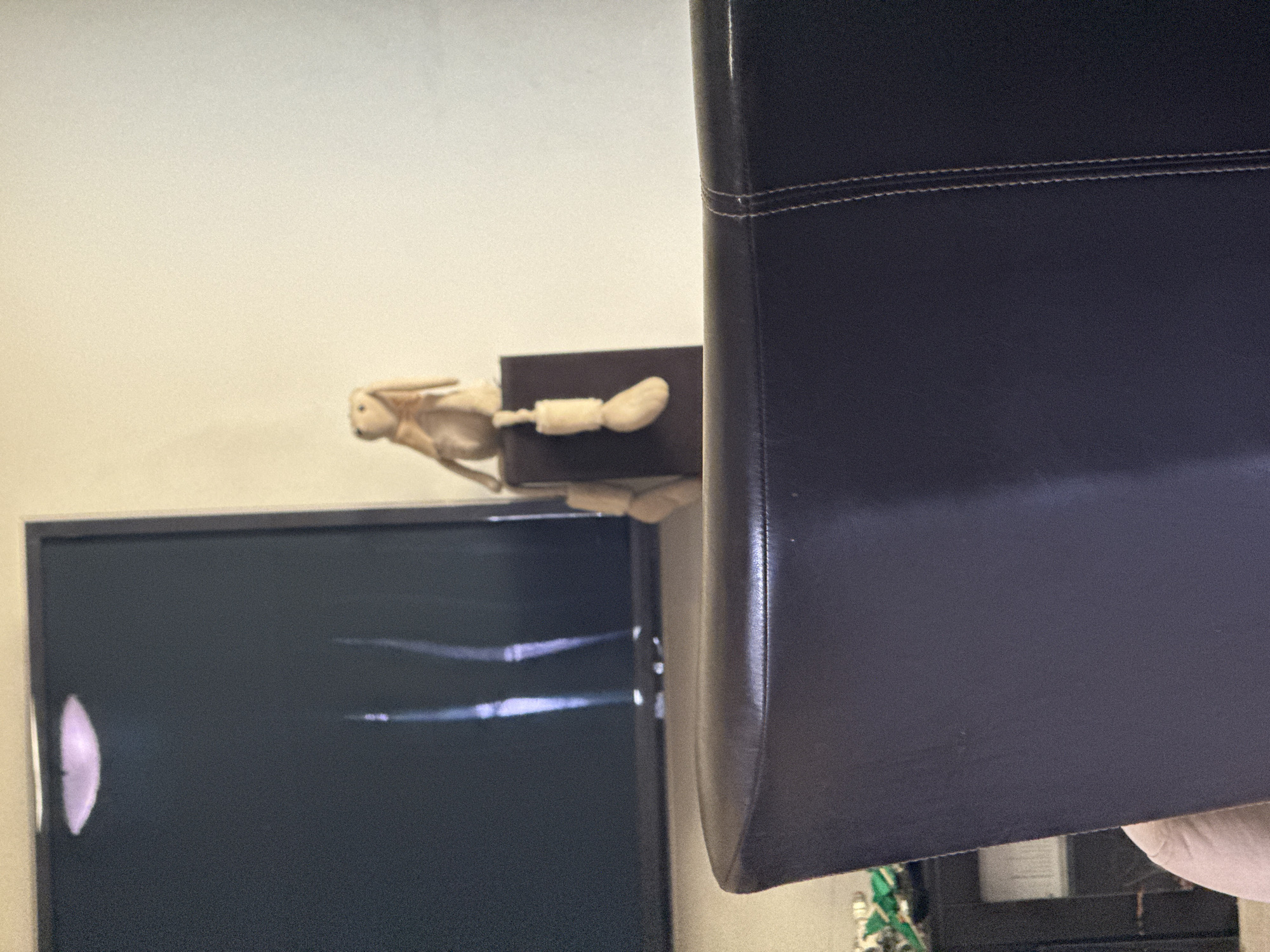 In a residential room in Broadbeach, Australia, a brown plush toy rabbit with long ears sits perched on a dark brown cube, its wooden legs dangling over the side. The rabbit is positioned to the right of a large, dark television screen displaying a faint, abstract light pattern. In the foreground, the back of a dark brown leather chair, with visible white stitching, occupies the lower portion of the frame, obscuring the lower half of the scene. The wall behind the rabbit and television is a neutral cream color.