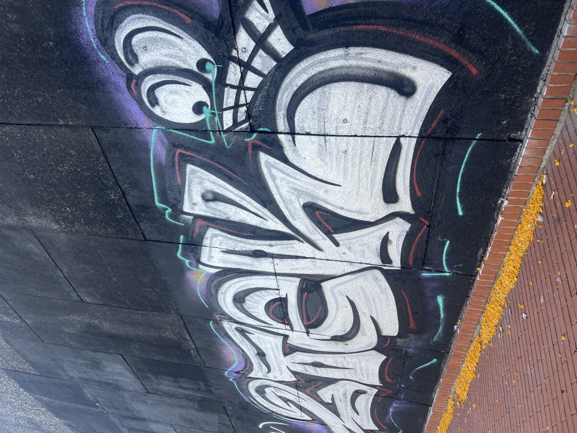 The image shows a section of a black concrete wall with graffiti art. The graffiti features large, stylized white lettering with black and red outlines, and a cartoonish face with large eyes and a gaping mouth. The wall appears to be outdoors, possibly a retaining wall, as suggested by the brick paving at its base. Scattered yellow flower petals are visible on the brick paving. The graffiti is vibrant and appears to be a recent addition to the wall. The scene is set in a public area, likely a street or walkway, in Barcelona, Spain.