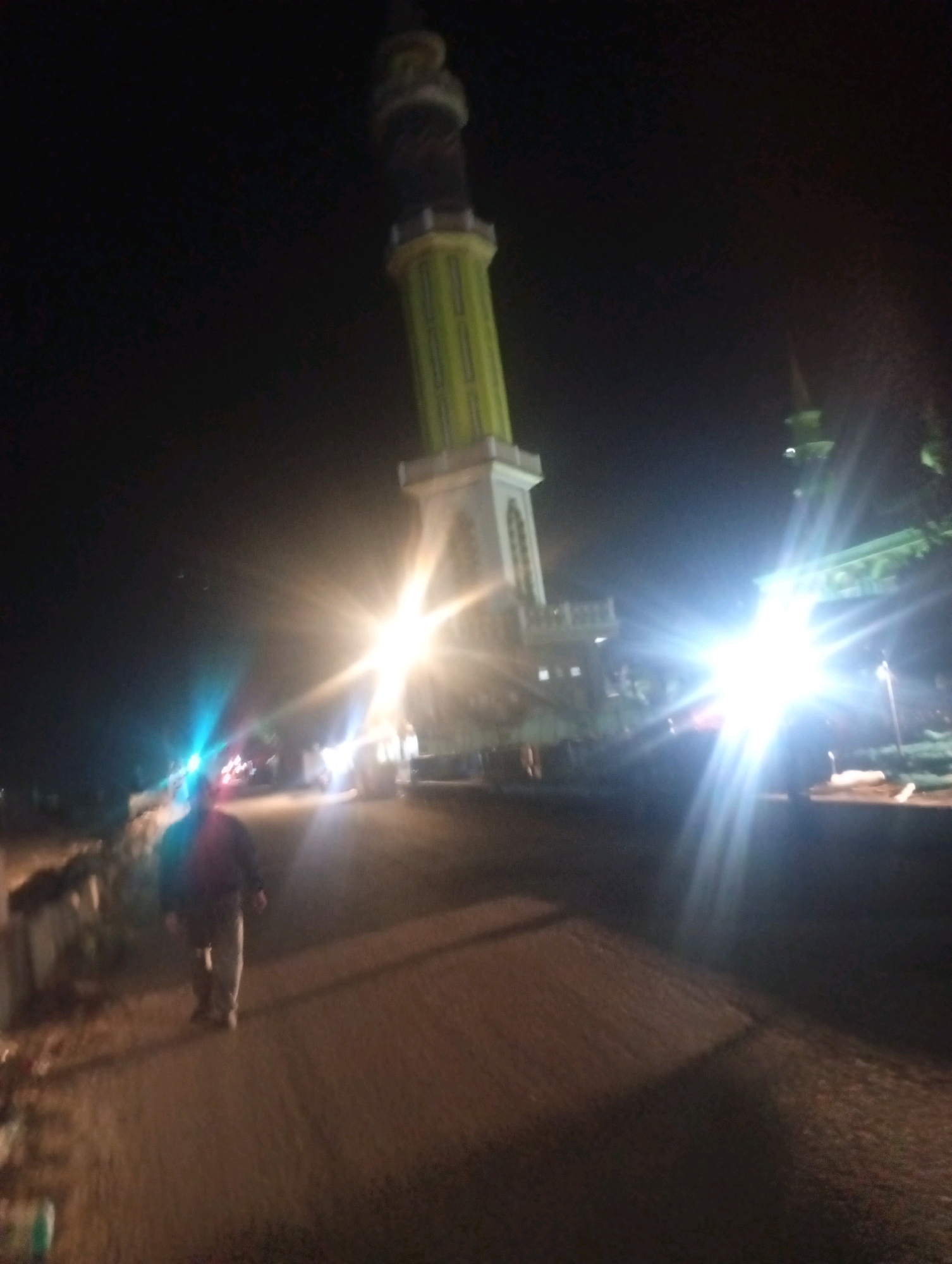 A person walks on a dirt road at night in front of a mosque with a prominent green and white minaret in Bajingjowo, Indonesia. The scene is illuminated by bright lights, possibly from the mosque or nearby vehicles, casting long shadows and creating lens flare effects. The overall image is dark and slightly blurry, suggesting it was taken in low-light conditions.