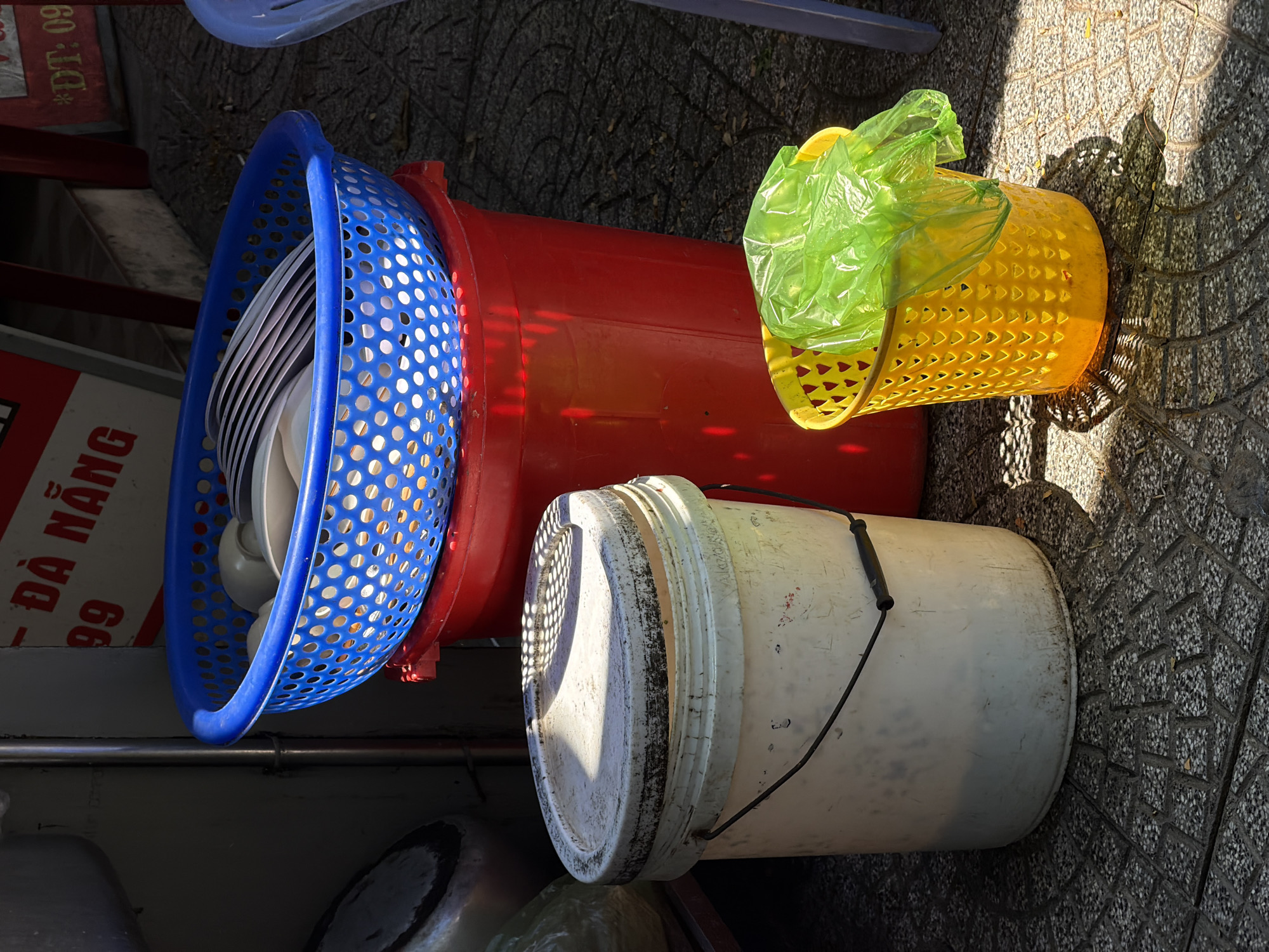 In Đà Nẵng, Vietnam, the image shows a collection of plastic containers on a patterned sidewalk. A blue plastic basket filled with plates and bowls rests against a larger, red plastic container. Beside the red container, a yellow plastic basket is partially covered with a green plastic bag. Adjacent to these, a white plastic bucket with a metal handle sits on the sidewalk. The scene appears to be an outdoor storage area, possibly associated with a nearby business, as suggested by the sign partially visible in the background.