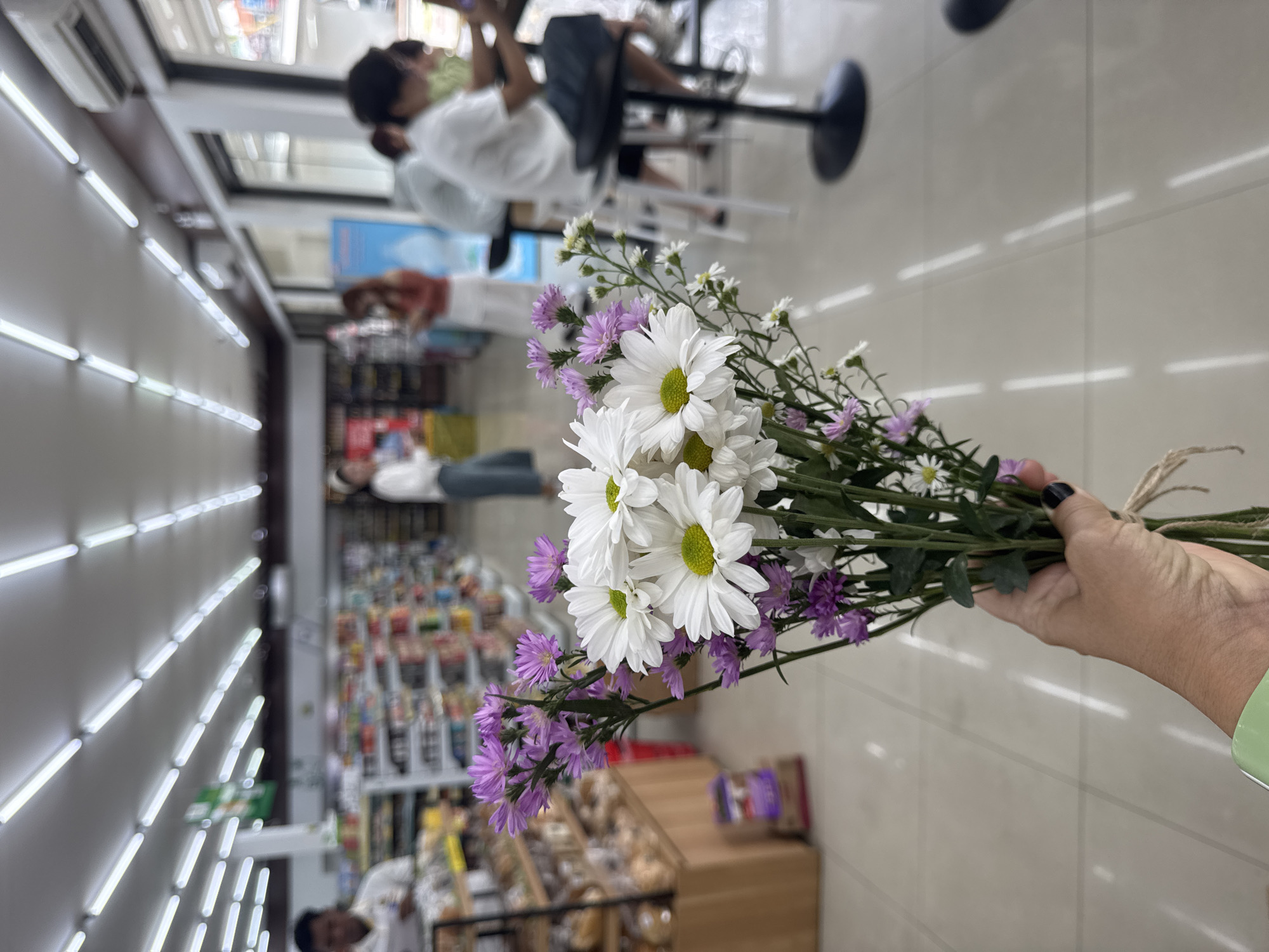 A hand holds a bouquet of white daisies and purple asters in the foreground. Behind the flowers, the interior of a convenience store is visible. The store has bright fluorescent lighting on the ceiling and shelves stocked with various products. Several people are present in the store, some standing and browsing, others seated at tables or counters. One person in the background appears to be an employee, wearing a white shirt. The floor is tiled. The scene suggests a typical retail environment. The image was captured in Kuta Selatan, Indonesia.