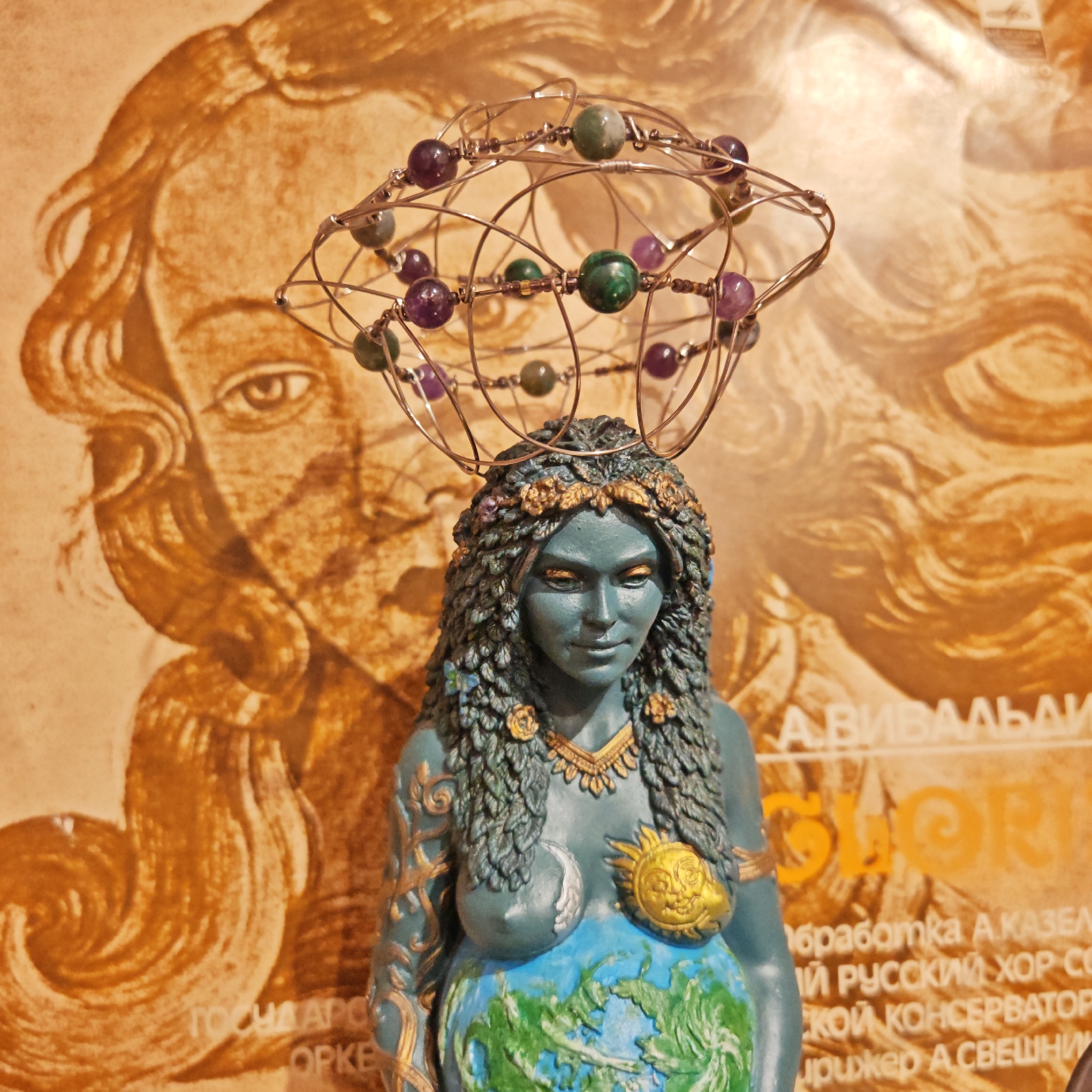 The image shows a statue of a female figure with blue skin, wearing a headdress made of wire and stones. The statue is positioned in front of a poster featuring a drawing of a woman's face. The statue has a necklace and arm bands, with a sun and moon symbol on her chest and the earth painted on her stomach. The poster behind it has Russian text on it, including the words "А.ВИВАЛЬДИ" (A. Vivaldi) and mentions orchestra and a choir from a conservatory. The picture appears to have been taken in Rostov, Russia.