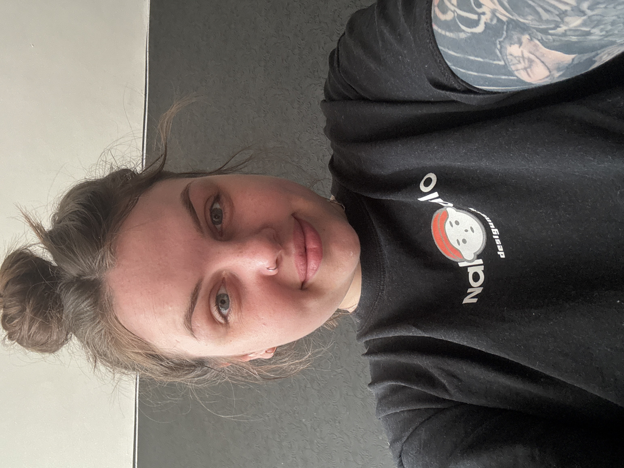 A person is looking at the camera. They have their hair in a bun, and have a nose piercing. The person is wearing a black shirt with a graphic on it. There is an arm with a tattoo visible. The background is gray and white. There is no information about the city of Chornomorskoe, Ukraine in the image.