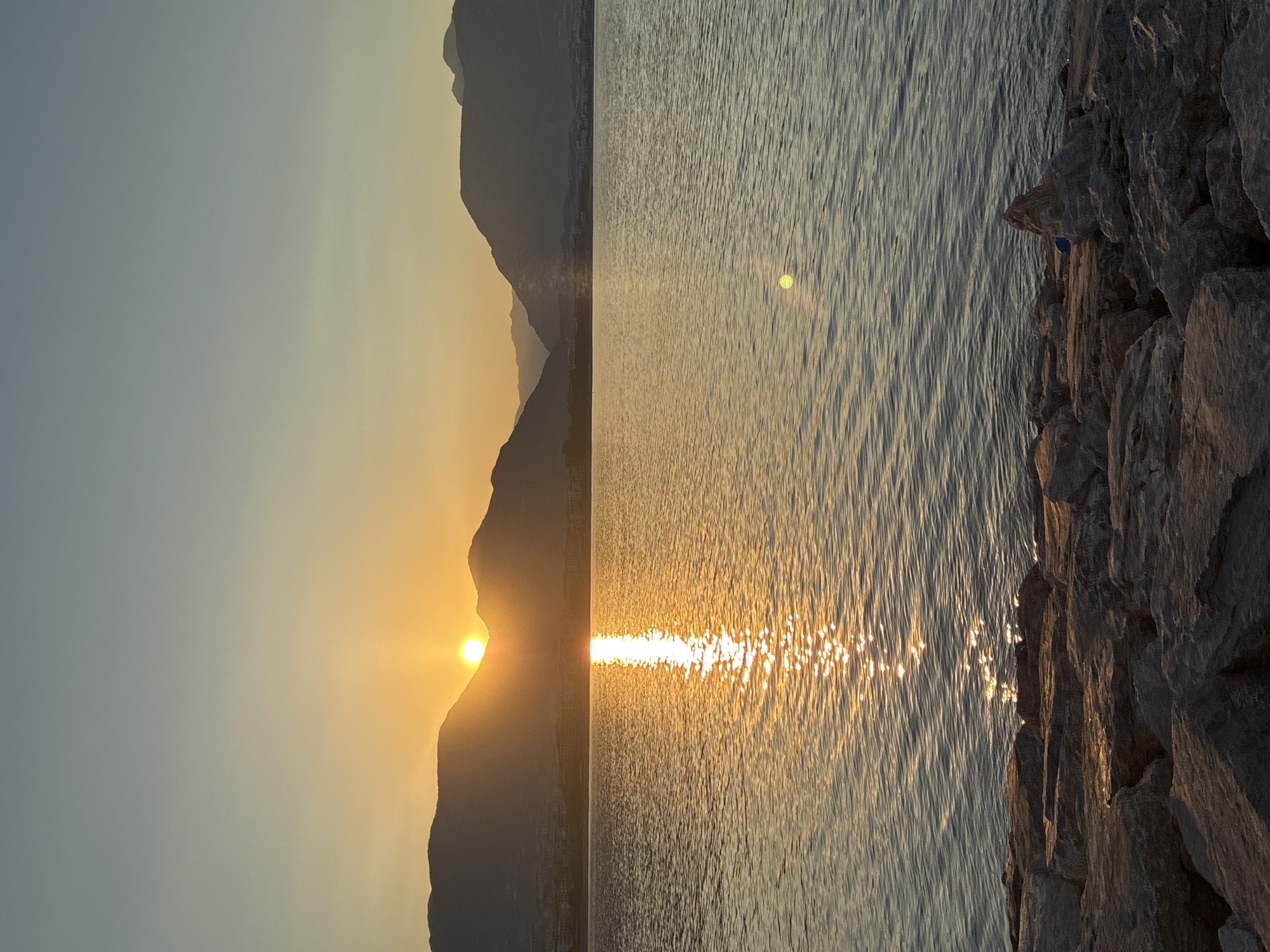 A sunset is captured over a body of water, likely the Mediterranean Sea, in Alanya, Turkey. The sun, a bright orb, is partially obscured by the silhouette of a mountain range in the background, casting a warm golden glow across the water's surface. The reflection of the sun creates a shimmering path on the rippling water. In the foreground, large, weathered rocks form a rocky shoreline. The sky transitions from a pale blue at the top to a soft orange and yellow gradient near the horizon. No people are visible in the image.