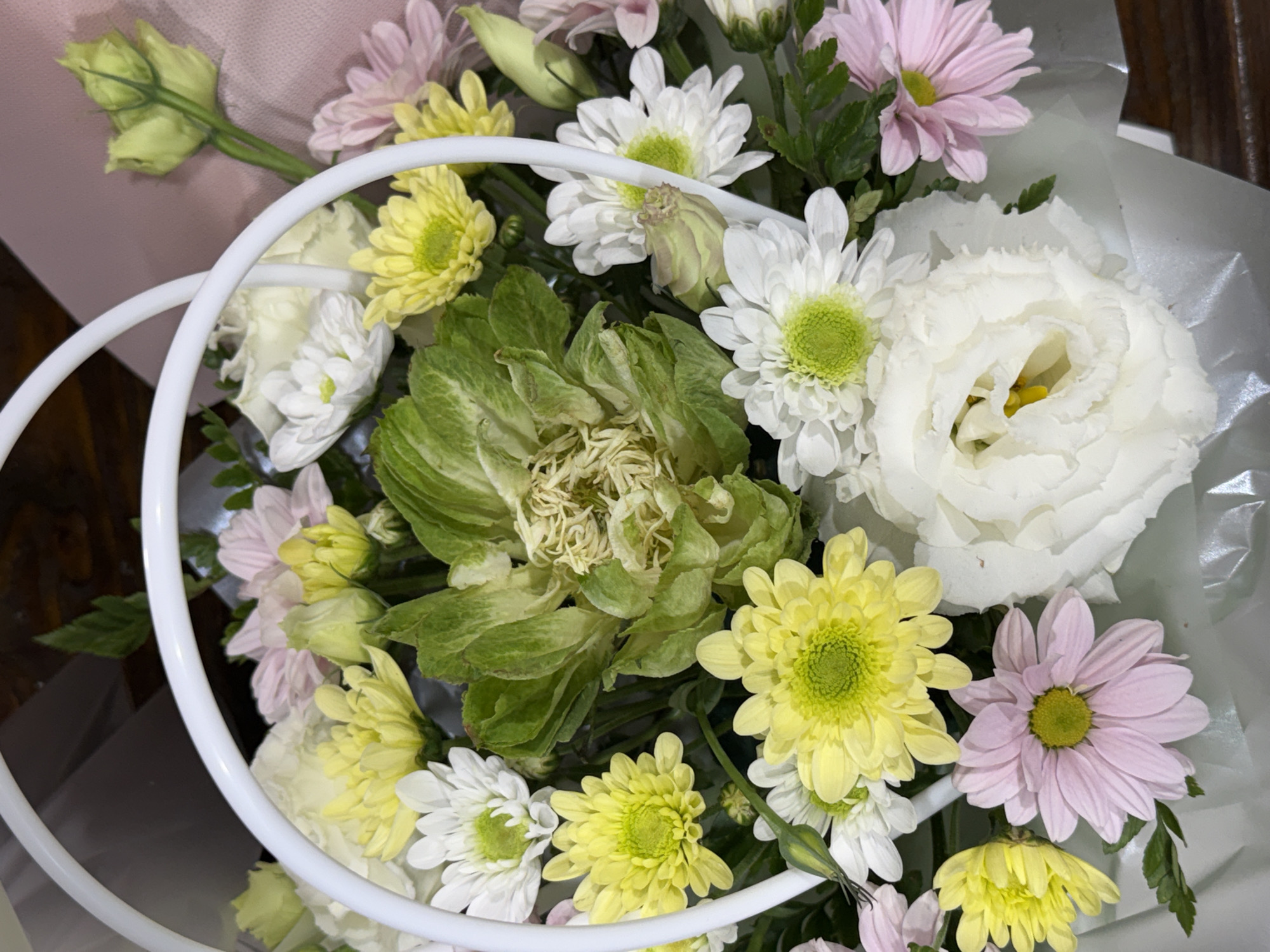A floral arrangement is presented in a white wire basket. The arrangement includes a variety of flowers, such as yellow and pink chrysanthemums, white daisies, and a large white rose. A distinctive green element, resembling lettuce or a tightly furled cabbage, is situated in the center of the arrangement. The backdrop is blurred, suggesting an indoor setting, possibly a table or countertop.
