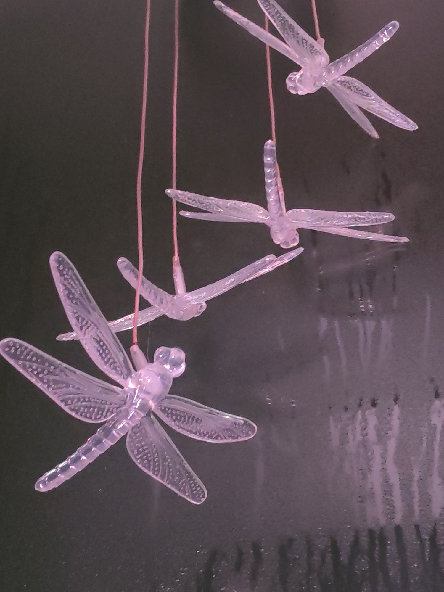 The image depicts a mobile of four translucent, pale purple dragonfly decorations hanging from thin, pink strings. The dragonflies are arranged vertically, decreasing in size from bottom to top. The background is a dark, mottled surface with streaks of a lighter purple hue, possibly a wall with textured finish or a reflective surface. There are no people visible. The location, Izhevsk, Russia, is not discernable from the image itself.