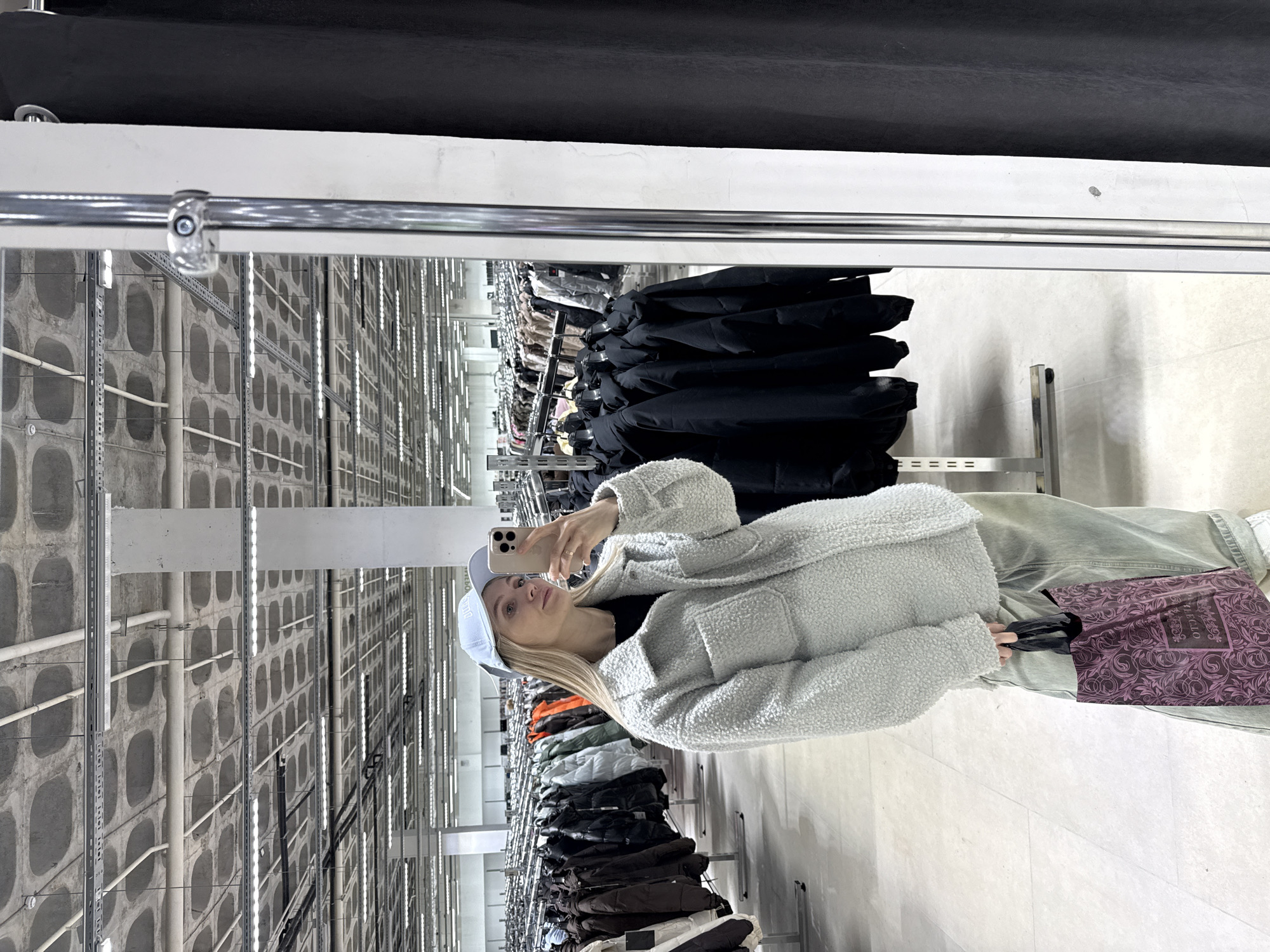 The image shows a woman taking a selfie in a clothing store. The woman is wearing a light blue baseball cap, a light gray, textured jacket, and light green pants. She is holding a shopping bag with a dark pink and purple pattern.
The store appears to be large, with rows of clothing racks extending into the distance. The walls are a light gray color, and the ceiling has a complex arrangement of pipes, lights, and structural supports. The clothing racks are filled with a variety of garments, including black jackets and clothing in various colors.
The store is located in Izhevsk, Russia.