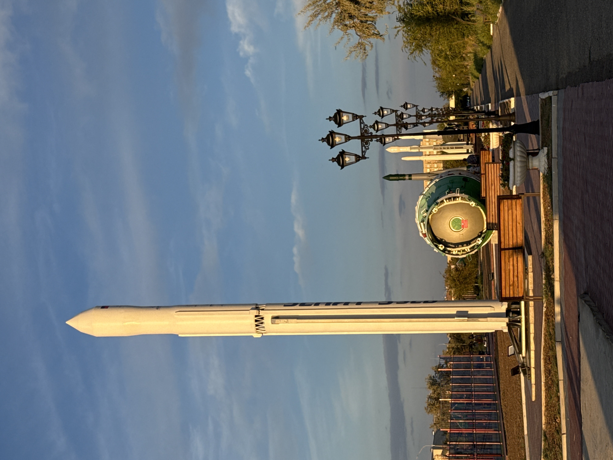 The image depicts an outdoor display of rockets and space-related artifacts in the city of Baikonur, Kazakhstan. In the foreground, a tall, white rocket stands vertically, dominating the left side of the frame. To its right, a large, circular, green and silver object, possibly a spacecraft or engine component, is displayed horizontally. Several other, smaller rockets are visible behind these primary exhibits.
A pathway made of red brick pavers leads into the scene, with wooden benches placed along its edge. Black lampposts with multiple ornate fixtures line the pathway, casting long shadows across the ground due to the low angle of the sun. The environment appears to be a park or public space, with grassy areas, trees, and a fence in the background. The sky is blue with scattered clouds, suggesting it is daytime. A few indistinct figures are seated on the benches. The scene is illuminated by the warm light of what appears to be either sunrise or sunset.
