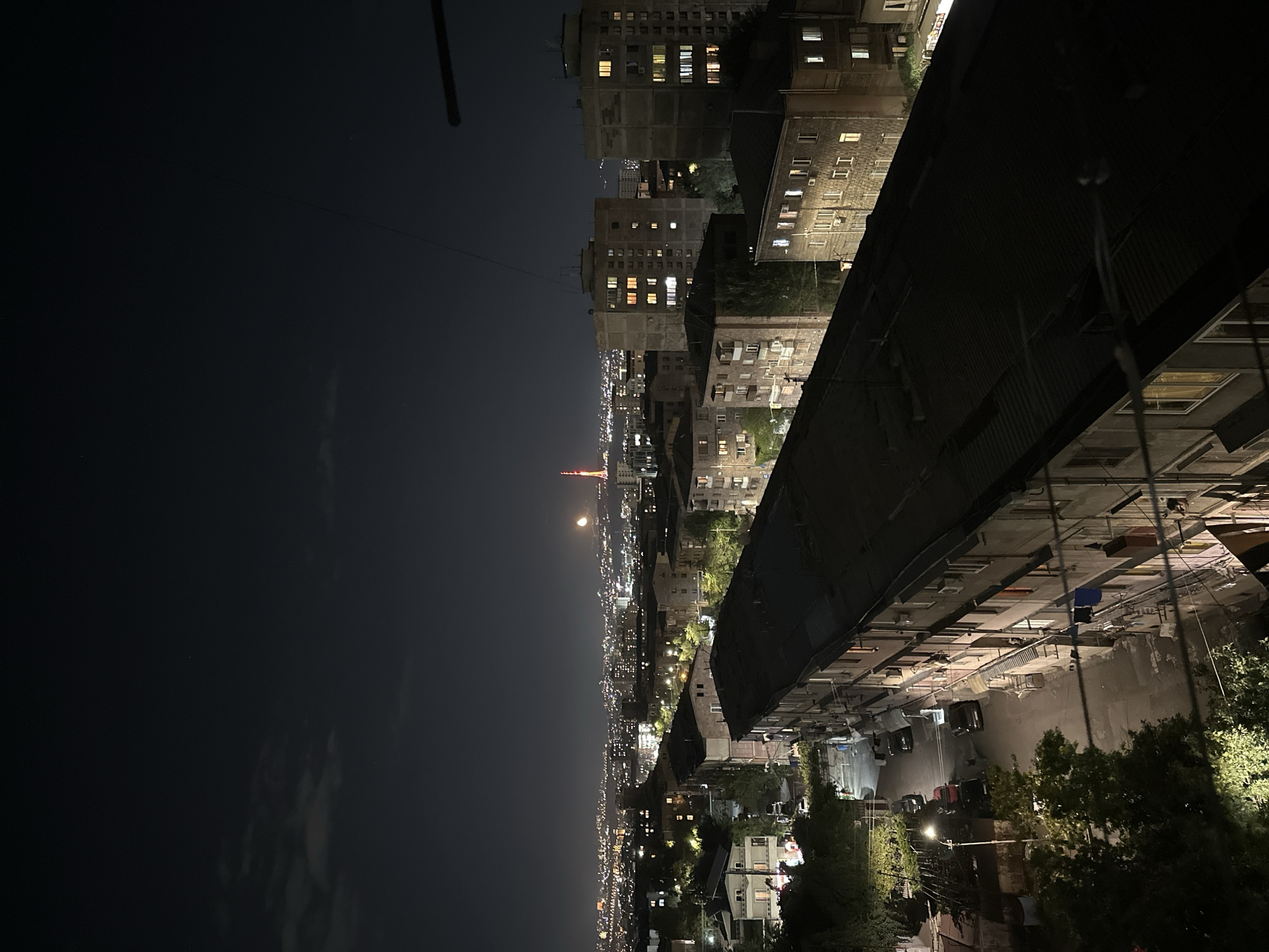 This image depicts a nighttime view of a residential area in Yerevan, Armenia. In the foreground, several multi-story apartment buildings, characteristic of Soviet-era architecture, are visible with lights on in some windows. The roofs of these buildings are dark and appear to be made of corrugated metal or similar material. A narrow street runs between some of the buildings, with several parked cars illuminated by streetlights and light from the buildings.
In the middle ground, more apartment buildings and trees are visible. Beyond this, the city of Yerevan stretches out, appearing as a vast expanse of lights. In the distance, a prominent landmark, the Yerevan TV Tower, is visible and illuminated. A full or nearly full moon is also visible in the sky, positioned to the left of the TV tower. The sky is dark with a few scattered clouds. The overall scene suggests a typical urban night in Yerevan, characterized by apartment living and the glow of city lights.