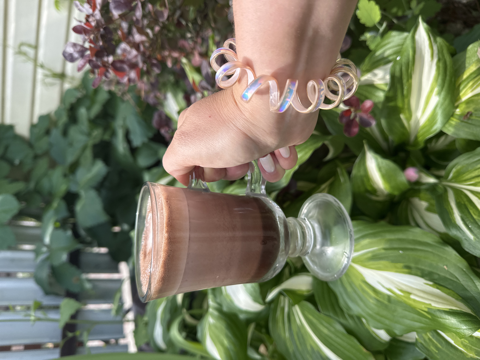 An image shows a hand holding a clear glass with a layered brown beverage, possibly hot chocolate or iced coffee. The hand is adorned with a spiral-shaped, iridescent bracelet. The background is a lush garden with large green and white variegated leaves and some dark purple foliage. A white wooden fence is visible in the far background. There is no information to suggest the location is in Nizhny Novgorod, Russia.