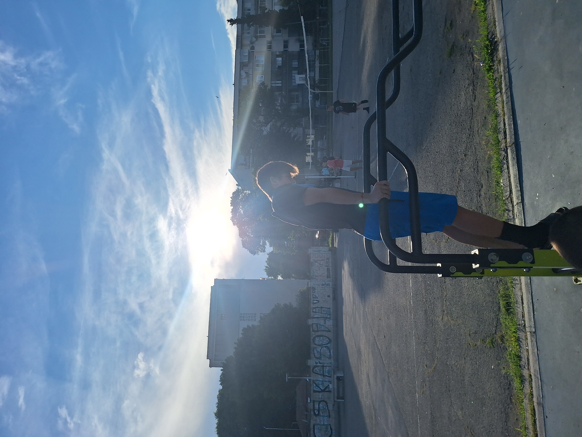 The image depicts an outdoor fitness area on a sunny day, likely in Varna, Bulgaria, given the context. In the foreground, a young person, silhouetted against the sun, is performing exercises on parallel bars. They are wearing blue shorts and a dark t-shirt. The fitness equipment is black with green accents. In the background, several apartment buildings are visible. A volleyball net is set up, and several individuals are playing. One person in a red shirt and white shorts is visible on the volleyball court, as are two other figures in dark clothing. Graffiti covers a low wall in the mid-ground. The sky is blue with scattered white clouds, and the sun is shining brightly, creating lens flare. The ground is paved, with some patches of grass and dirt visible. The overall scene suggests a recreational area within a residential neighborhood.