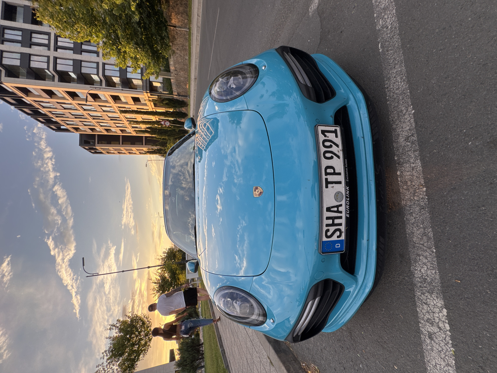 A bright blue Porsche Panamera is parked on asphalt, angled towards the camera. The car's front is prominent, showing its distinctive headlights and front bumper. A license plate is visible on the front, with the text "SHA TP 991" and "EUROLiNK Exclusive Autos". To the left of the car, a modern building with many windows stands, reflecting the warm glow of the setting sun. The sky is filled with dramatic clouds, illuminated by the golden hour light, suggesting late afternoon or early evening. Two people are standing near the car, their backs to the camera, enjoying the view or a conversation. The scene appears to be in an urban or suburban setting, possibly a parking lot or street near a residential area in Burgas, Bulgaria. The overall mood is peaceful and serene, amplified by the beautiful sunset.