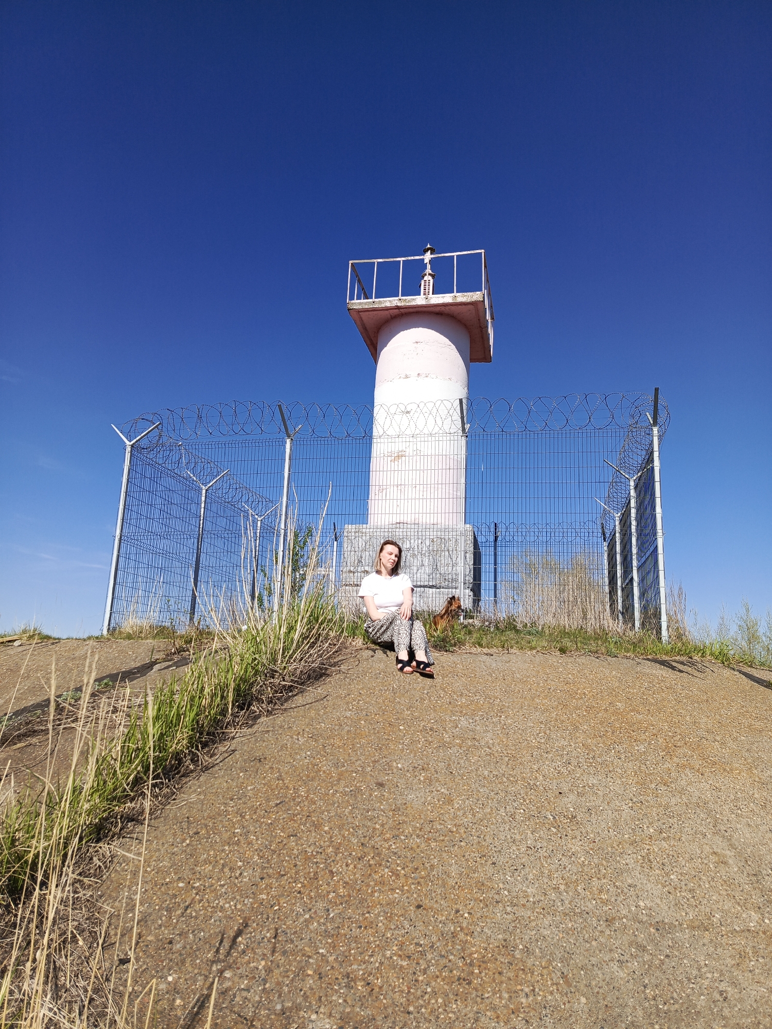 A woman sits on a dirt embankment in front of a white, cylindrical structure enclosed by a wire fence topped with barbed wire. She wears a white t-shirt, patterned pants, and sandals. A dog is partially visible to her right, behind the fence. Patches of tall grass grow along the embankment and near the fence. The sky is clear and blue. This location is likely near the city of Chaikovsky, Russia.