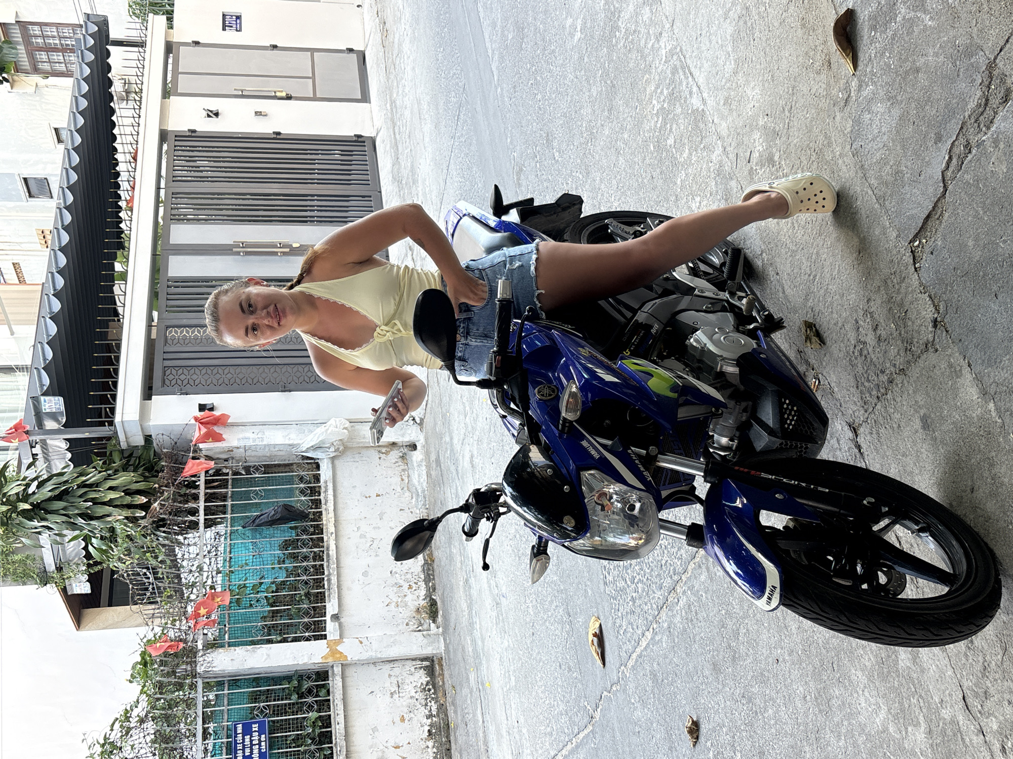 A woman with a braided hairstyle is posed sideways on a blue and black Yamaha motorcycle in Nha Trang Ward, Vietnam. She is wearing a yellow tank top and denim shorts, with white Crocs on her feet and holding a smartphone in her hand. The setting appears to be an urban street with a building featuring a gray gate in the background and Vietnamese flags visible. A sign indicating parking instructions is posted on the lower left, partially visible due to the orientation of the image.