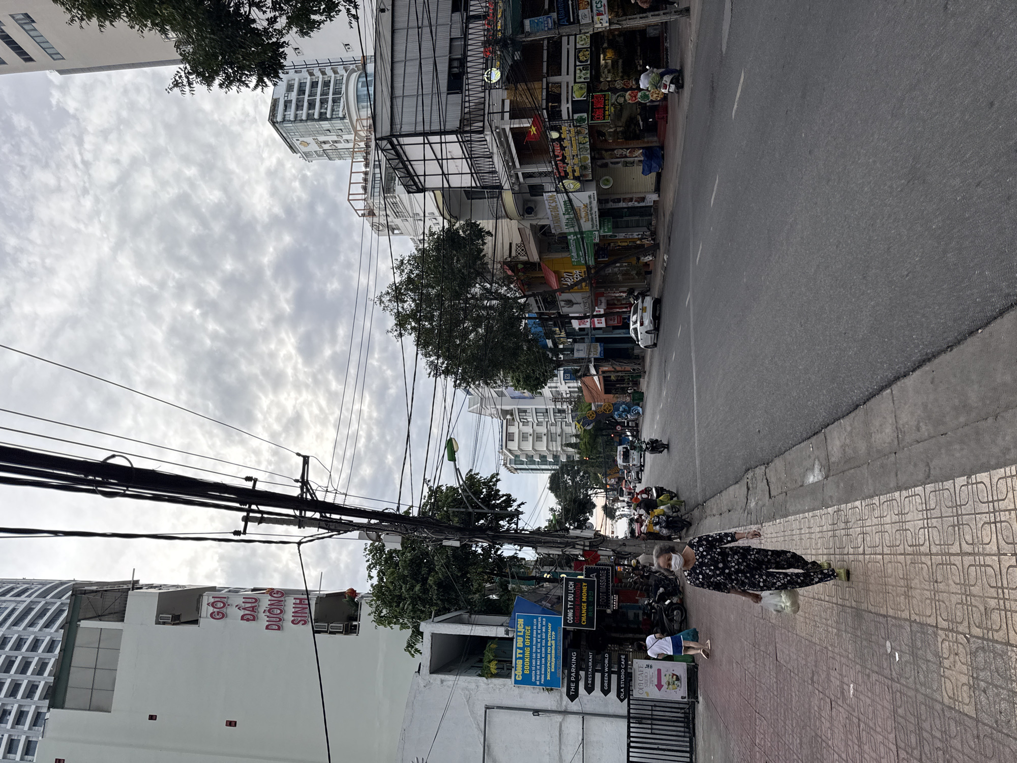 A street scene in Nha Trang, Vietnam, shows a paved road with white lane markings, flanked by sidewalks and multi-story buildings. Power lines crisscross above the street, supported by utility poles. On the left sidewalk, a woman wearing a floral-patterned outfit and a face mask walks towards the camera, carrying a plastic bag filled with white items. Further down the sidewalk, a younger person in a white shirt and blue shorts walks away from the camera. Several motorcycles are parked along the sidewalk, and a few are on the road. A white car is visible on the road in the distance. The buildings on the right side of the street feature various signs and awnings, indicating businesses such as a booking office, money exchange, and cafes. The sky is overcast with clouds.