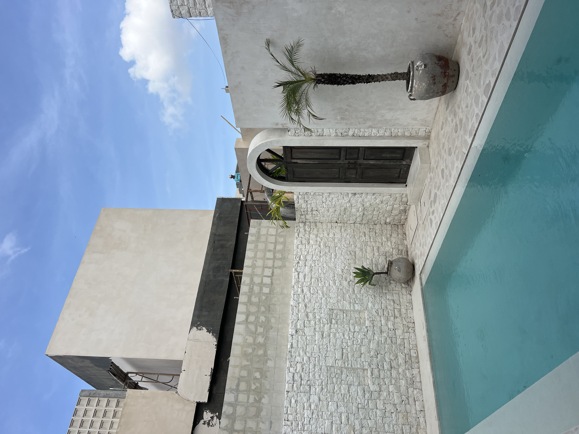 The image captures a contemporary architectural scene, most likely a luxurious villa, situated in North Kuta, Indonesia. The composition is dominated by the stark white facade of the building, accented by a dark wooden arched doorway and segments of exposed brickwork. The overall aesthetic evokes a minimalist and modern design.
A slender palm tree in a large pot adorns the upper wall, adding a tropical touch to the structure. Further down, another potted plant rests against a textured white brick wall near the edge of an azure-colored swimming pool. The pool's still water reflects the daylight.
In the background, the corner of a concrete building is visible, possibly a neighboring structure or a different section of the same property. Scaffolding is attached to the wall. The blue sky, adorned with sparse clouds, completes the backdrop of the scene.