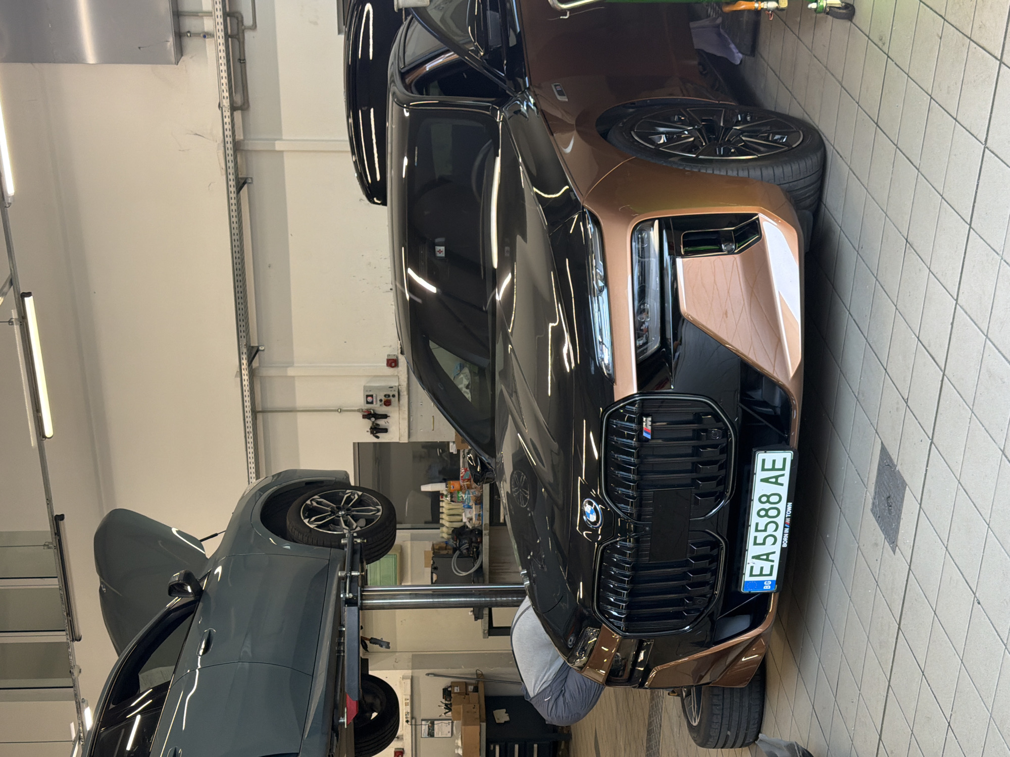 The image depicts a BMW 7 Series sedan, likely a G70 model, in a car workshop. The car is black with copper accents on its front bumper and side skirts. It has a Bulgarian license plate reading "BG EA 5588 AE." The car is positioned in the center of the frame, with its front end facing the viewer. A second car, a grey SUV with its hood open, is elevated on a car lift in the background to the left. A person wearing grey work pants and a grey shirt is bent over, working on the front of the BMW. The environment is a well-lit indoor workshop with tiled flooring and metal shelving units visible in the background, containing various car maintenance supplies. The presence of a Bulgarian license plate indicates the location is in Bulgaria. Burgas is a major city in Bulgaria, and this scene is consistent with a car service center within such a city.