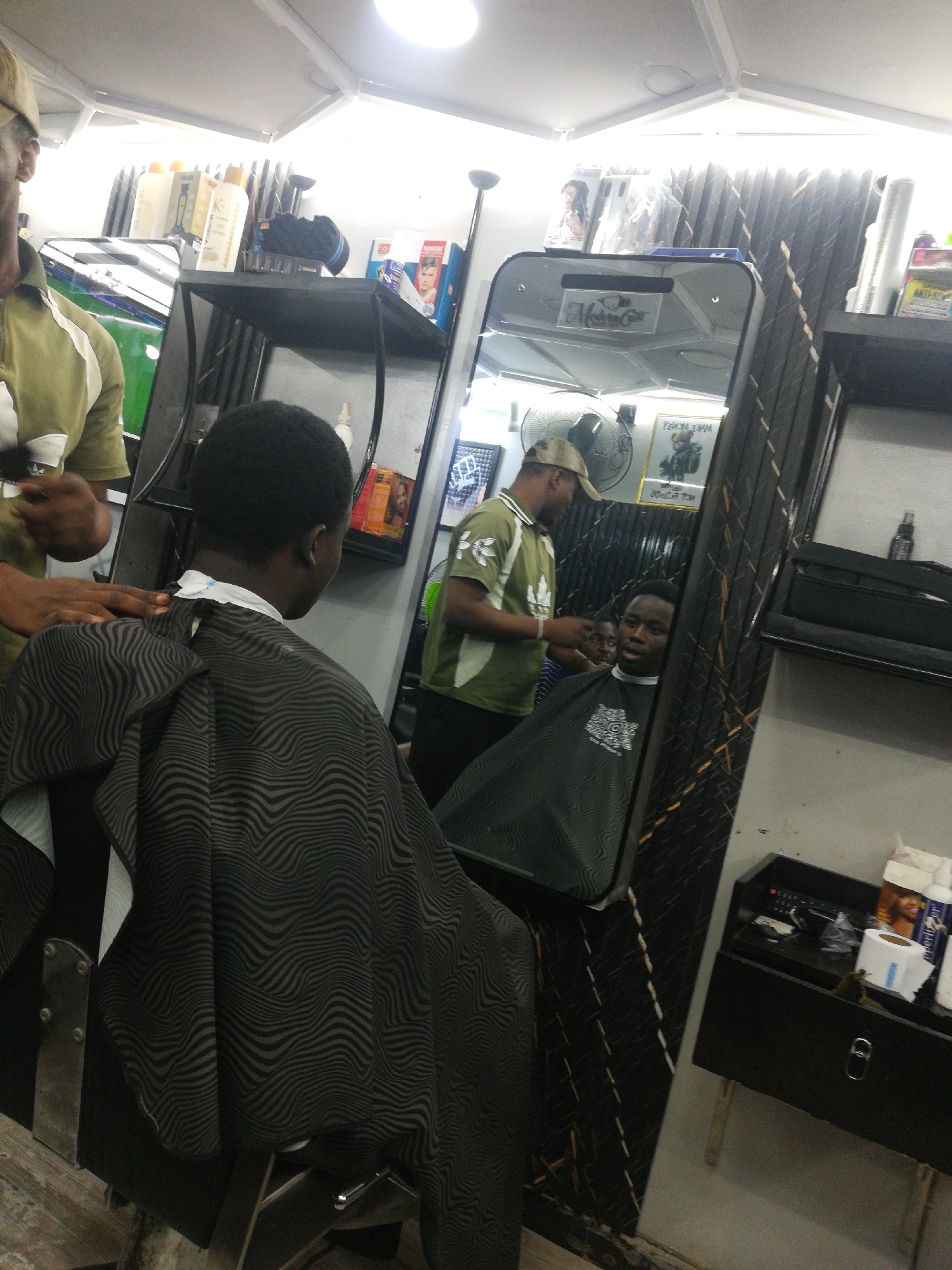 The image shows a scene inside a barbershop in Lagos, Nigeria. A young, dark-skinned man is seated in a barber's chair, draped with a black and gray striped cape. He faces a large, rectangular mirror. In the mirror's reflection, another dark-skinned man is visible, receiving a haircut from a barber. The barber wears a green shirt and a beige cap. Hairdressing tools and products are seen on shelves and a small black cabinet around the mirror. The lighting is bright.