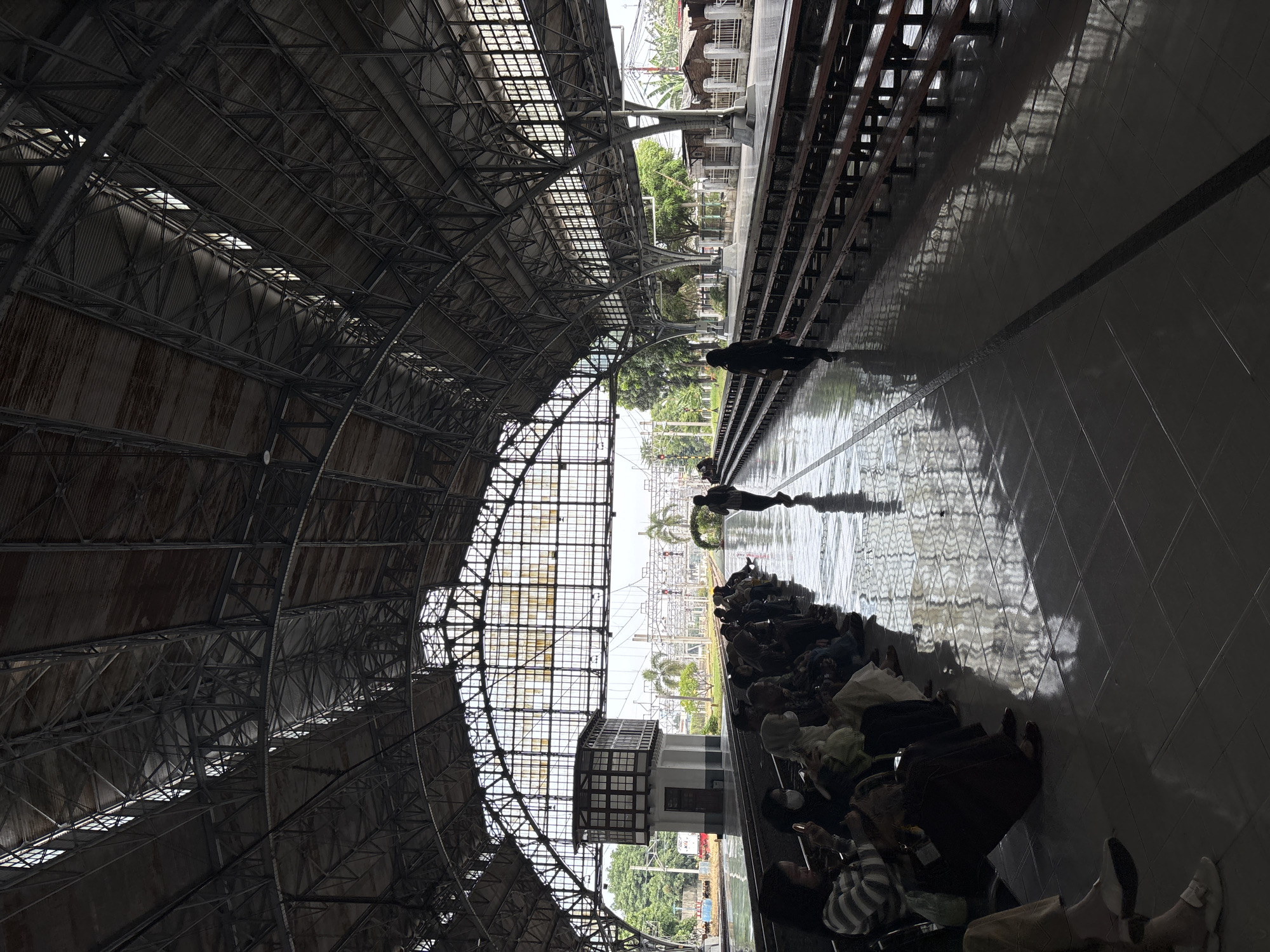 The image depicts a vast, arched train station platform under a high, metallic truss roof. The roof structure, composed of corrugated metal panels and numerous skylights, spans the upper portion of the frame. On the tiled platform to the right, a group of people is resting or waiting, some lying down or sitting on the floor with their luggage. Further down the platform, one person stands near the edge, and another is seen closer to the tracks.
Beyond the platform and tracks, a distinctive two-story building, likely a signal box, with a white base and a wooden, multi-windowed upper section, stands prominent. Outside the station, lush green trees and other buildings are visible against a bright, clear sky. The reflective tiled floor strongly mirrors the overhead structure and the bright light entering from outside.
The scene is illuminated by bright natural light, indicating daytime, possibly late morning or early afternoon, given the distinct shadows and reflections. The atmosphere suggests a period of waiting or transit. No visible text is present. The architectural style, including the large, industrial roof and the signal box, along with the outdoor vegetation, is consistent with a train station setting in North Jakarta, Indonesia.