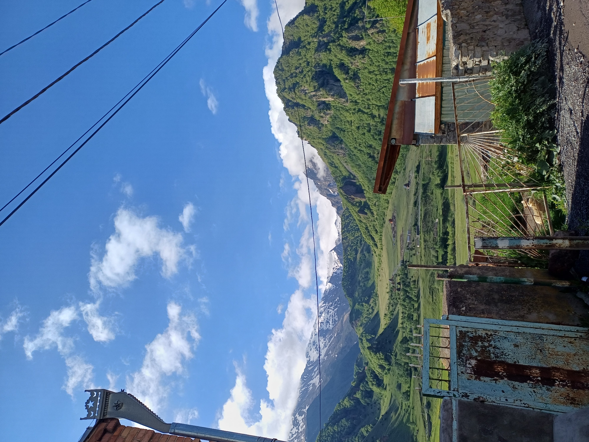 The image depicts a rural landscape with a prominent mountain range in the background. The mountains are a mix of green slopes and snow-capped peaks, indicating a high altitude. The sky is a vibrant blue with scattered white clouds. In the foreground, there are signs of human habitation, including a weathered blue gate and a rusty metal fence. A building with a corrugated metal roof is visible to the right, constructed with stone and metal walls. Beyond the fence, a green meadow stretches towards the mountains, dotted with small houses and trees. The overall scene suggests a serene, natural environment with a touch of rustic architecture. The location is identified as Stepantsminda, Georgia.