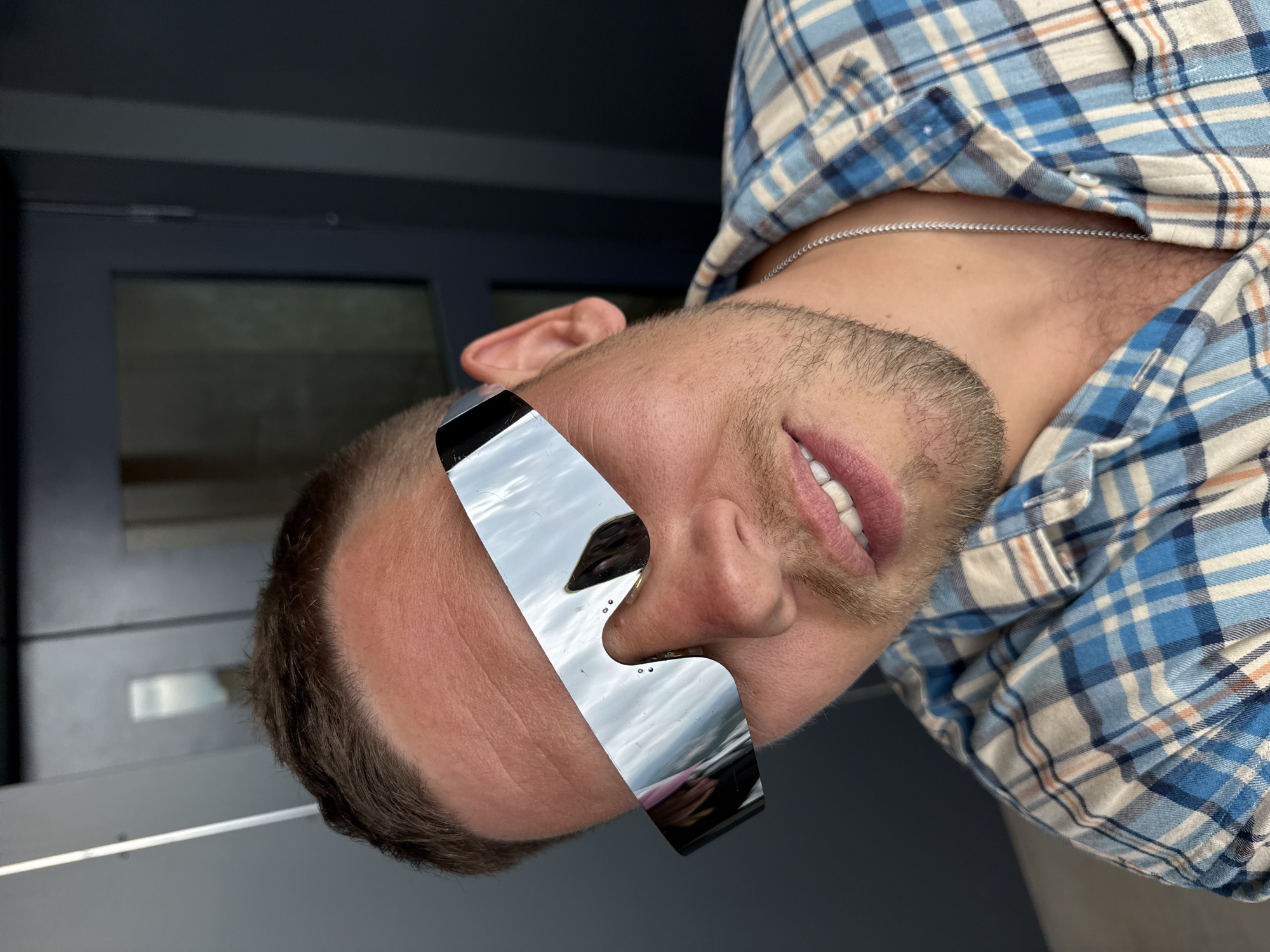 A person with short brown hair is wearing reflective sunglasses and a blue plaid shirt, angled with their head tilted to the left. A silver chain is visible around their neck. The background includes a gray surface with a window. There is no information that can identify the location as the city of Sochi, Russia.