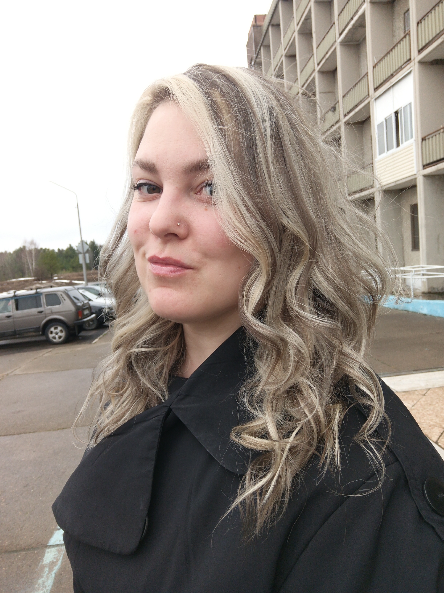 A woman is posing for a photo outside a building in Марковский, Russia. She has blonde hair with streaks and a nose piercing. She is wearing a black coat. The background includes parked cars, trees, and a multi-story building with balconies. The sky is overcast.