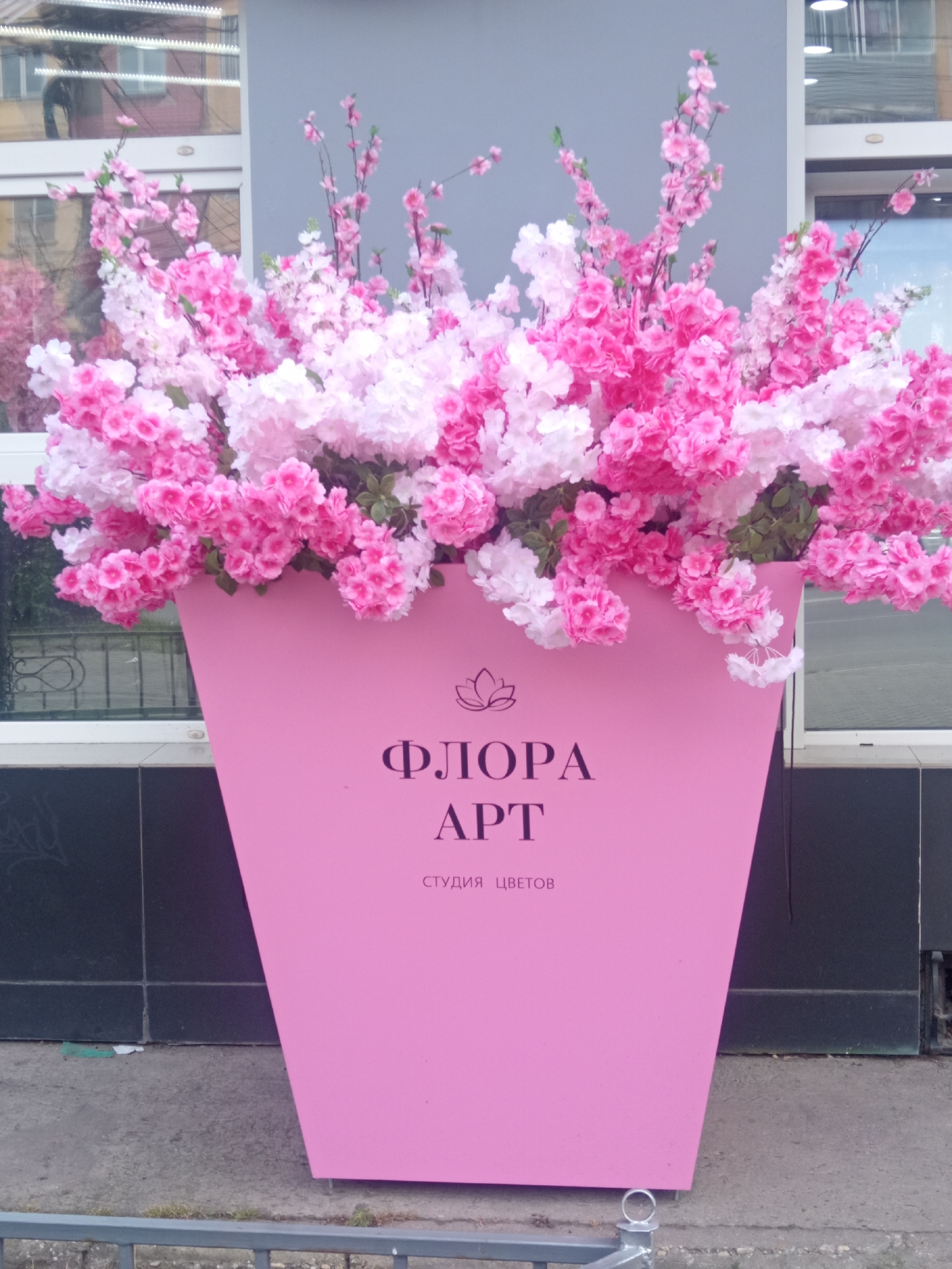 A large, bright pink planter is filled with artificial pink and white cherry blossoms. The planter has text in black, with a logo above it. The text reads "ФЛОРА АРТ" and below that "СТУДИЯ ЦВЕТОВ". The planter is positioned on a concrete sidewalk in front of a building with large windows that reflect the surroundings. The building appears to be commercial, with a gray facade and dark tiled lower section. A metal railing is visible in the foreground. The scene is set outdoors in the city of Belogorsk, Russia. There are no people or animals present in the image.